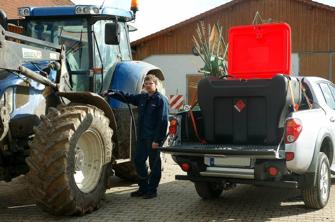 A man is standing between a blue tractor and a white pick-up truck with a fuel tank on its load bed. Agricultural buildings can be seen in the background.