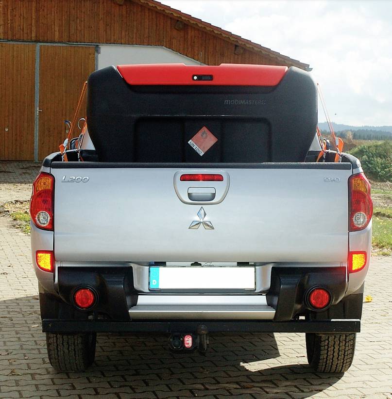 A silver pickup truck is parked in reverse in front of a wooden building. A large red engine sits in the truck's load bed.