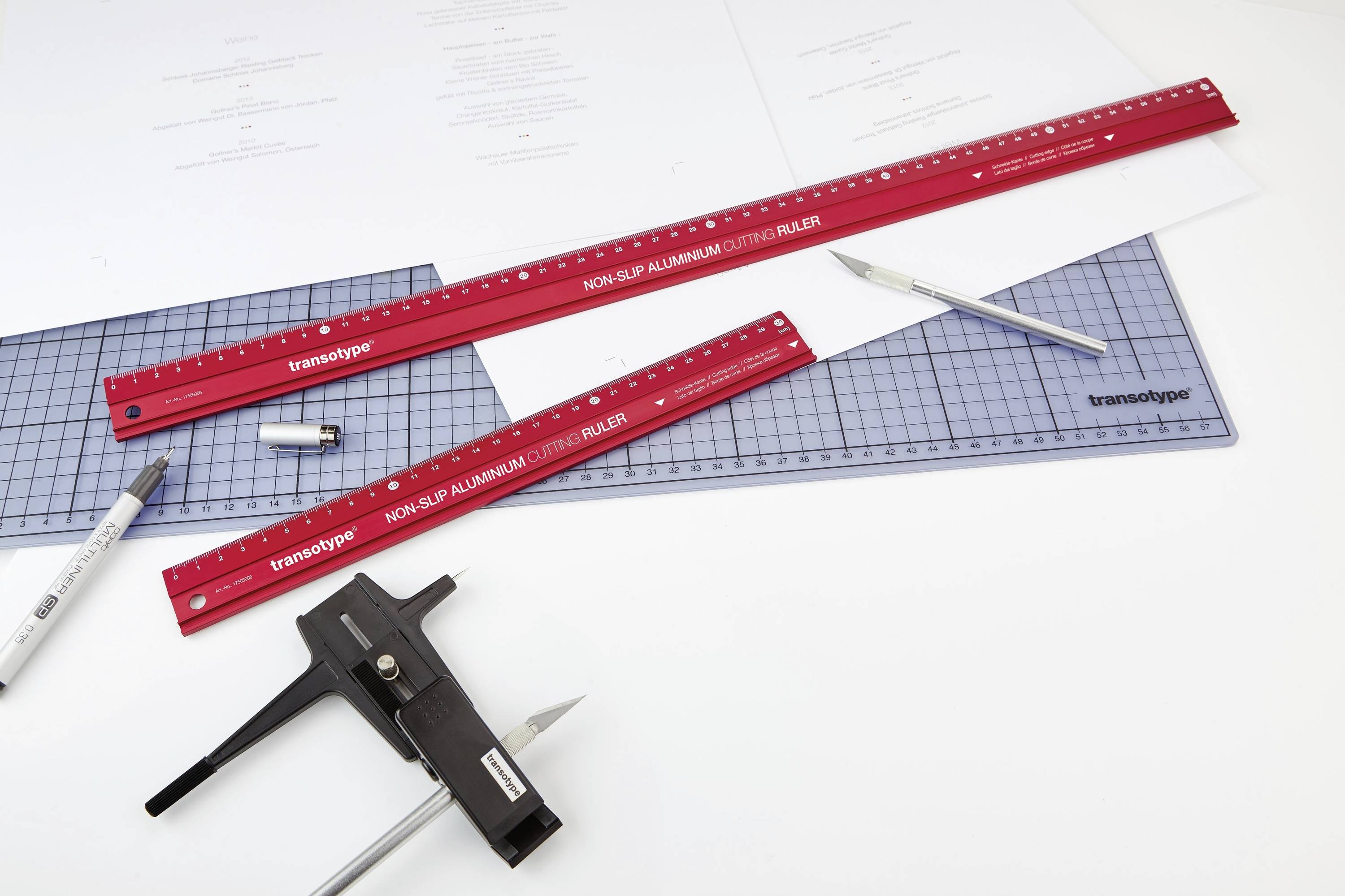 Two red rulers, a pen and additional drawing accessories are lying on a drawing board. The background shows drafts and ruler markings.
