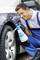 A man is cleaning a car tyre with a spray cleaner. He is wearing blue work trousers and a white polo shirt, and is sitting beside a car.
