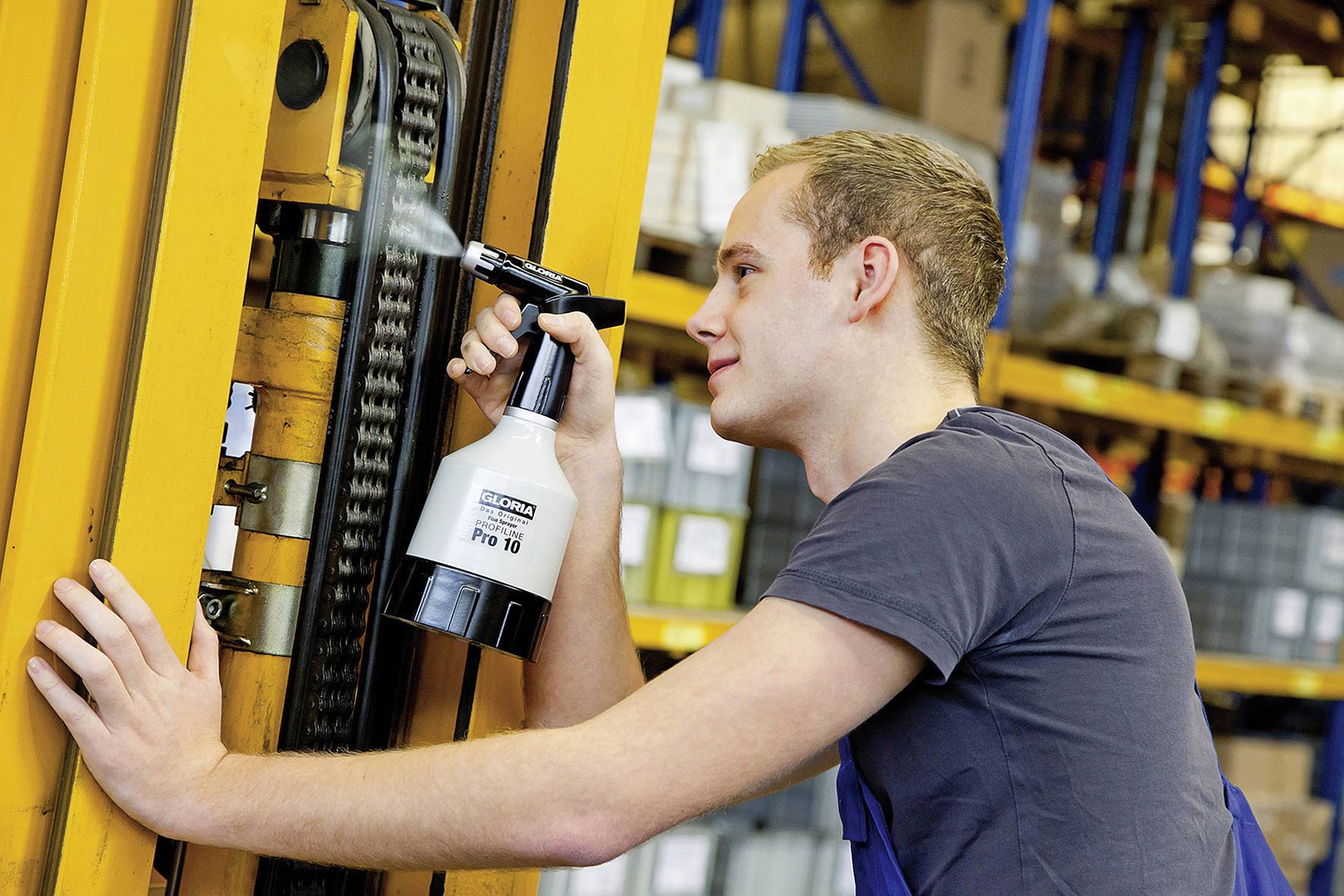 A man in a warehouse is maintaining a machine by spraying lubricant onto a chain. Shelves with cardboard boxes are visible in the background.