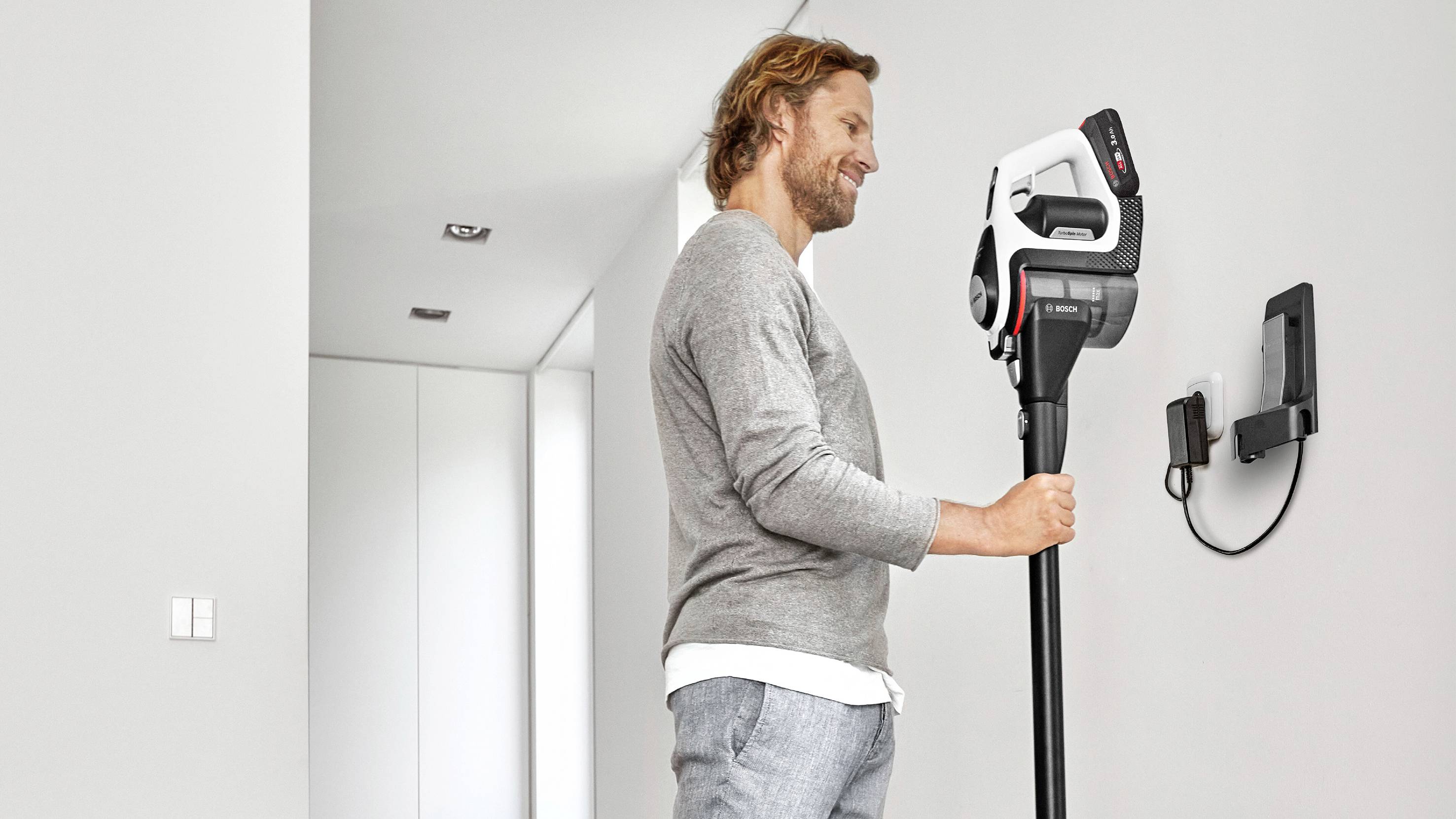 A man is using a cordless vacuum cleaner to mount a wall bracket. He is standing in a modern, bright room.