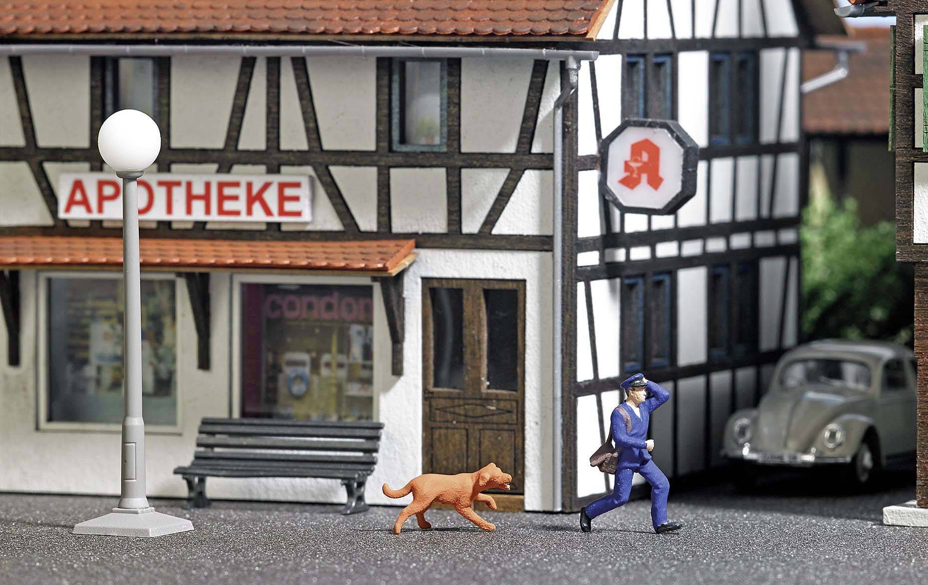 A miniature model village with a half-timbered building, marked by a pharmacy sign. A figure walks past with a dog.