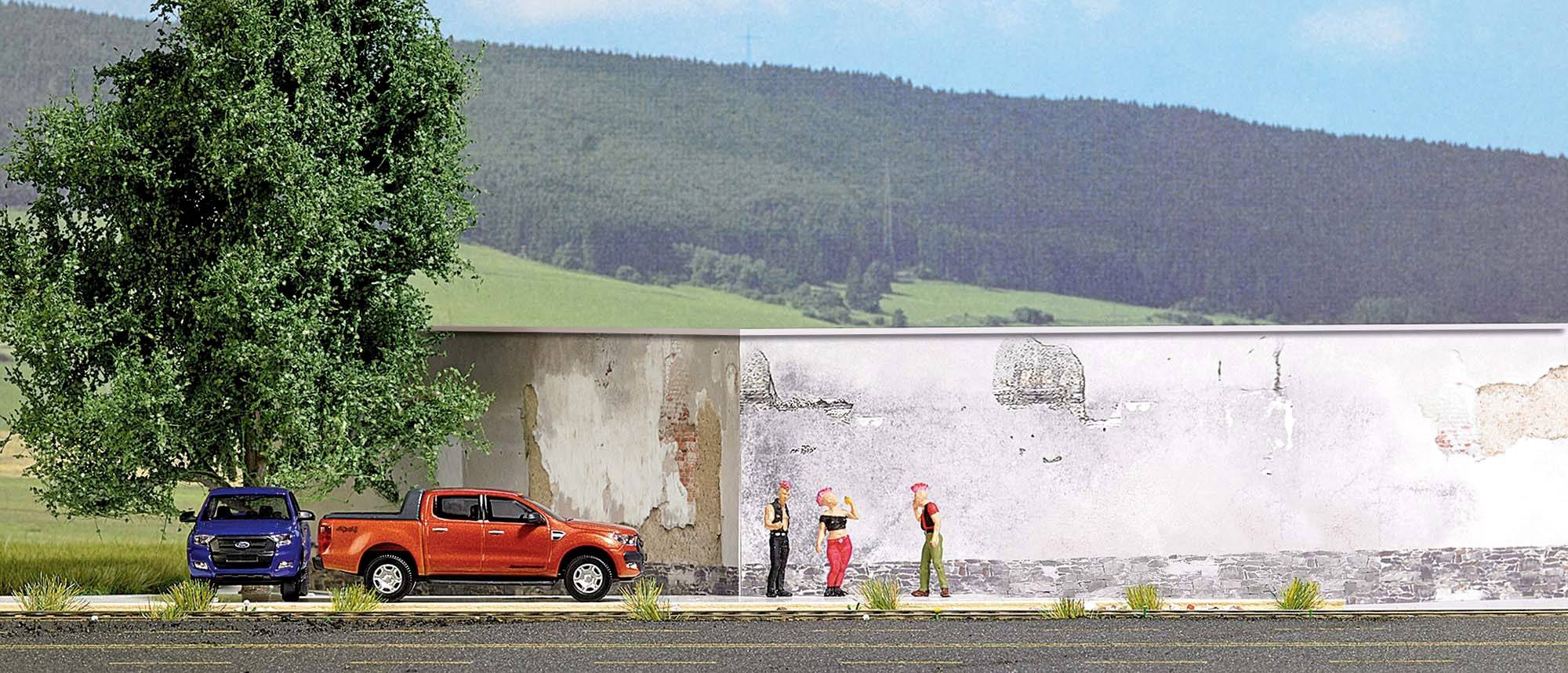 A red and a blue car are parked on a street. Three people are standing in front of a weathered wall, with a landscape in the background.