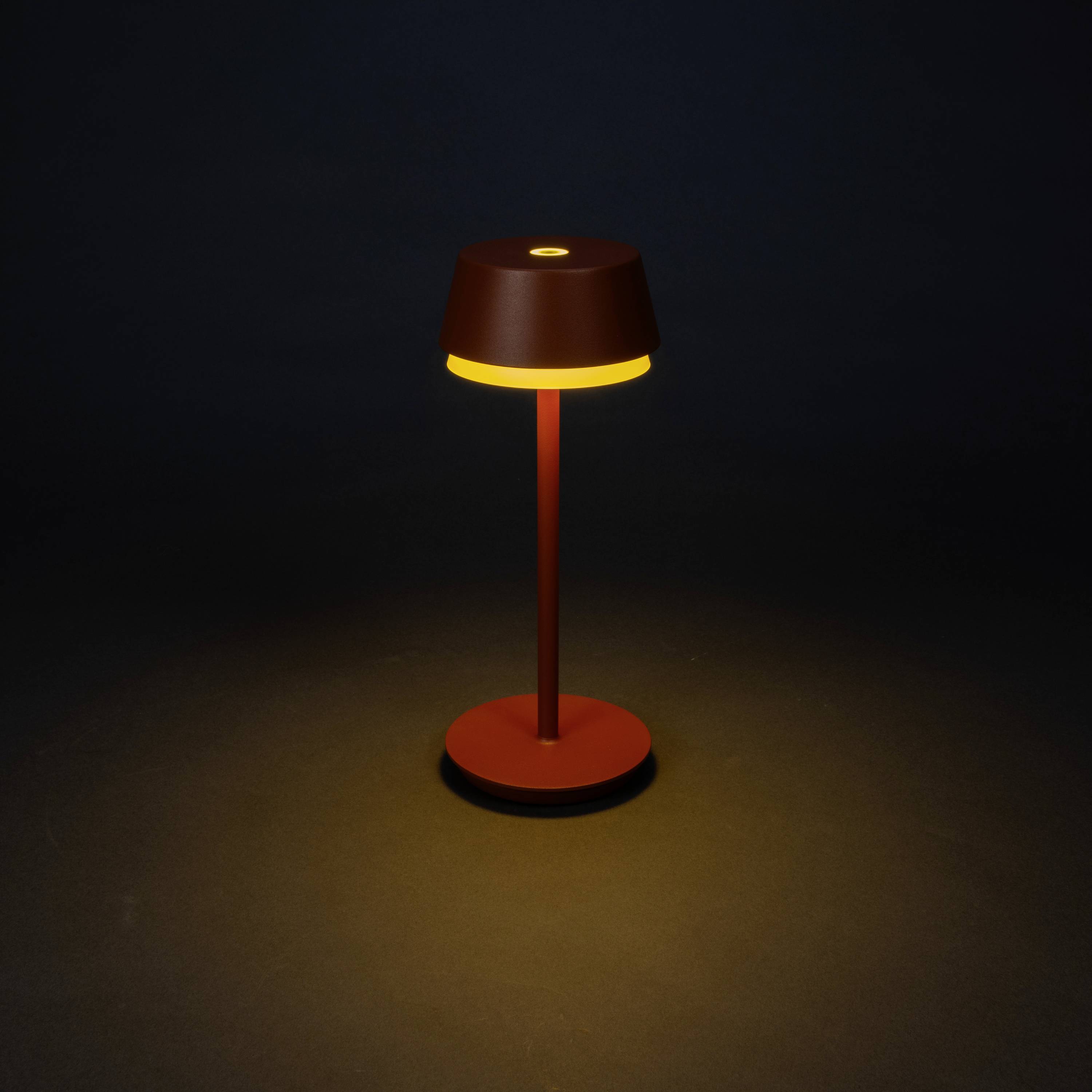 A modern table lamp with a brown shade emits warm yellow light, standing in a dark room.