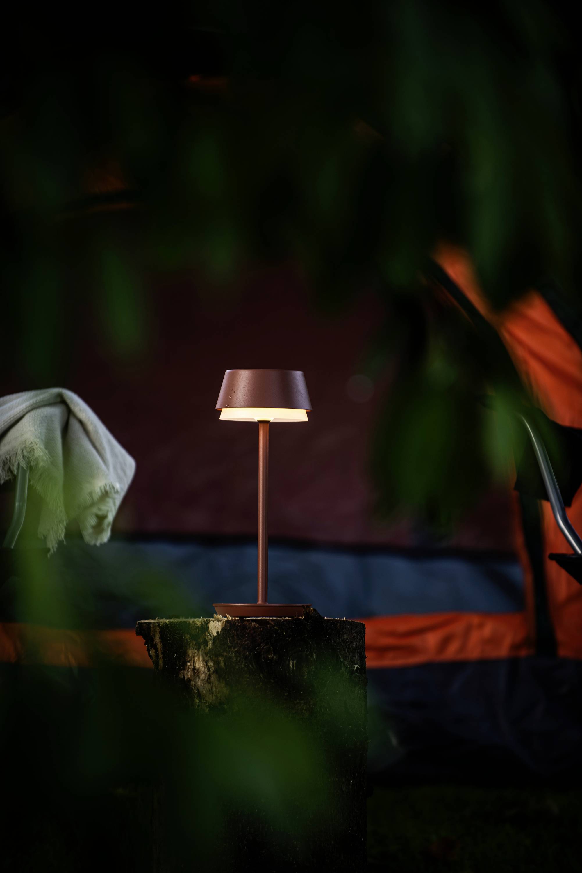 A table lamp stands on a tree stump in front of a tent outdoors.