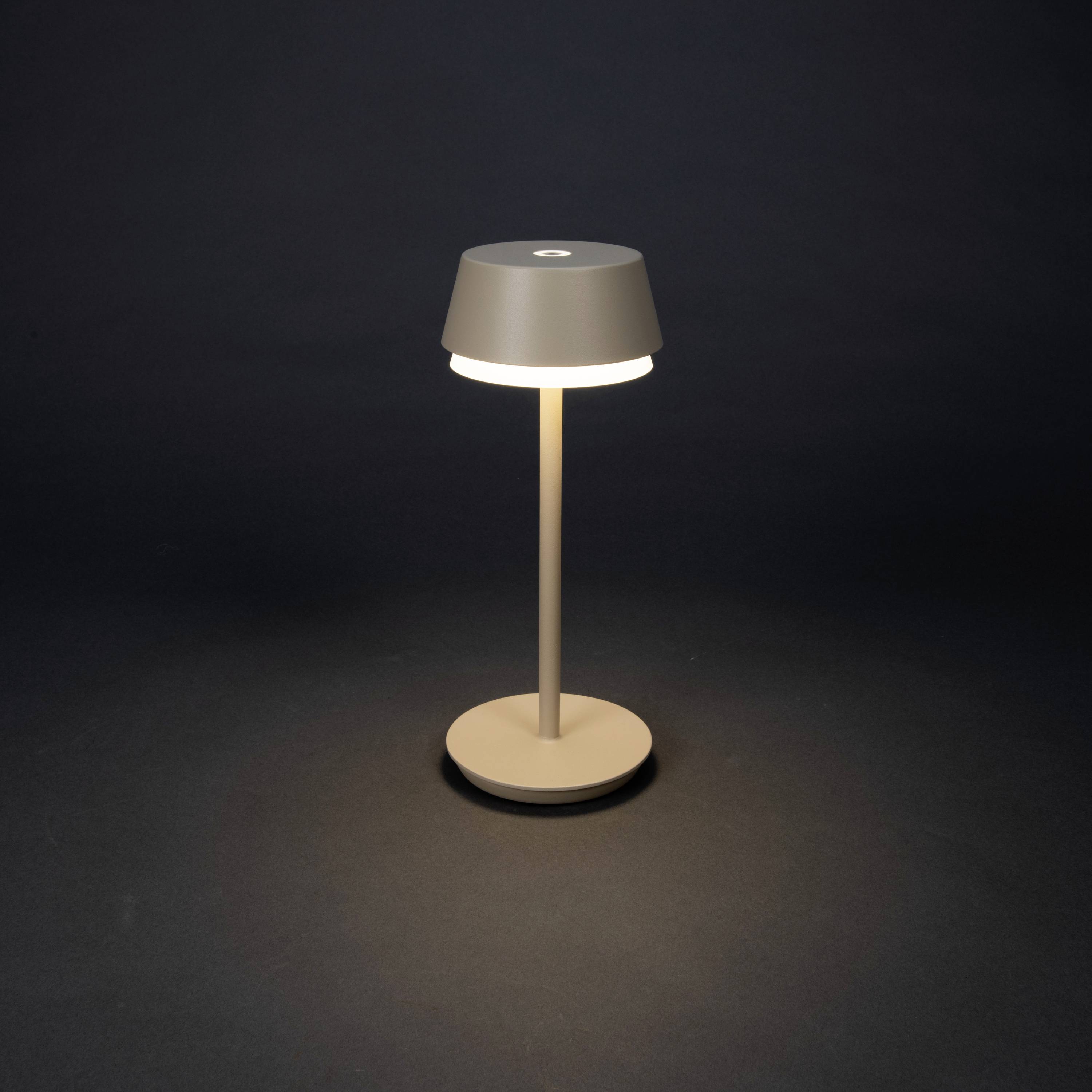 A modern table lamp with a simple, round lampshade illuminates a dark room and casts soft light across the surface.