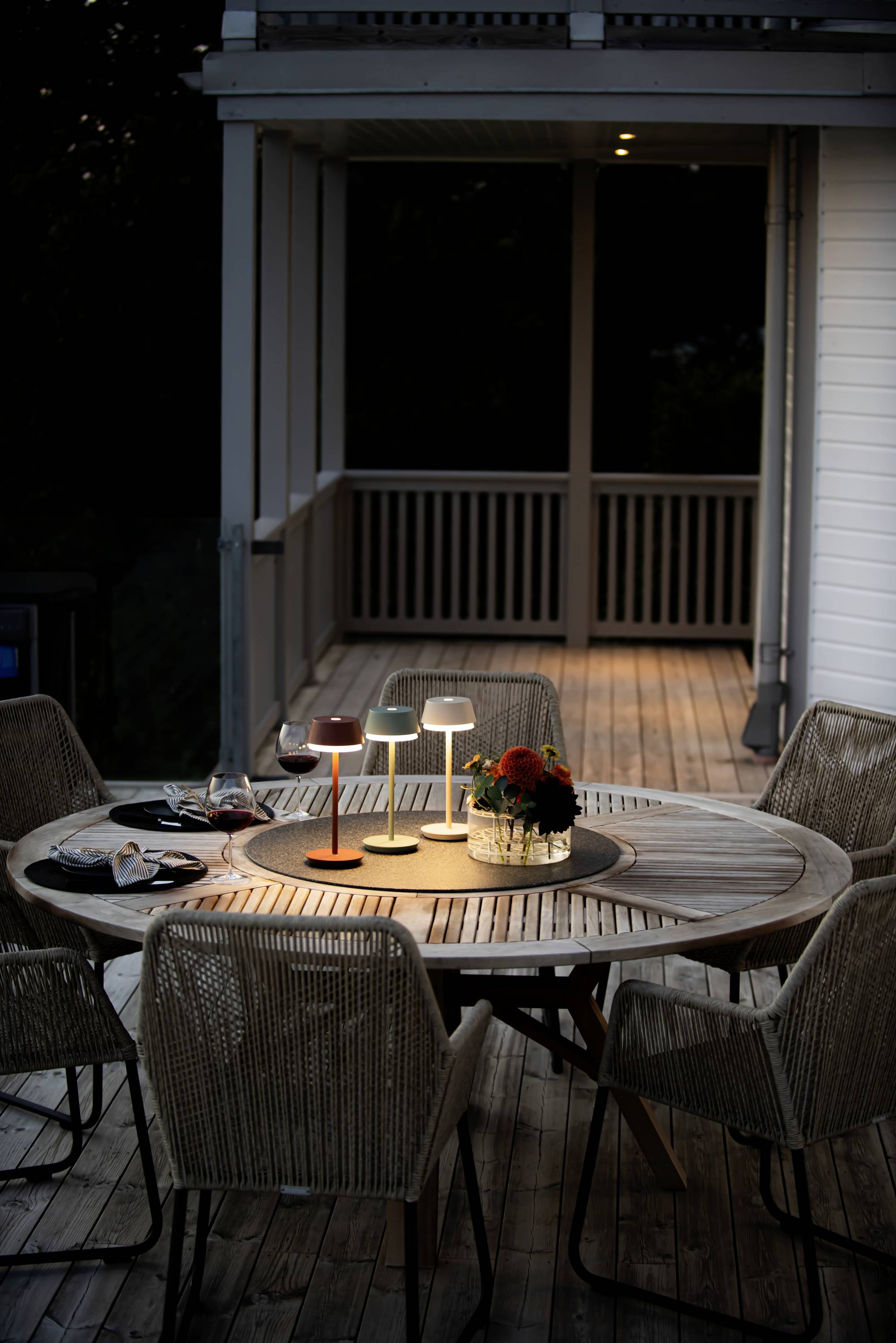 A round wooden table on a veranda, illuminated by three table lamps and decorated with flowers in a vase. Chairs surround the table.