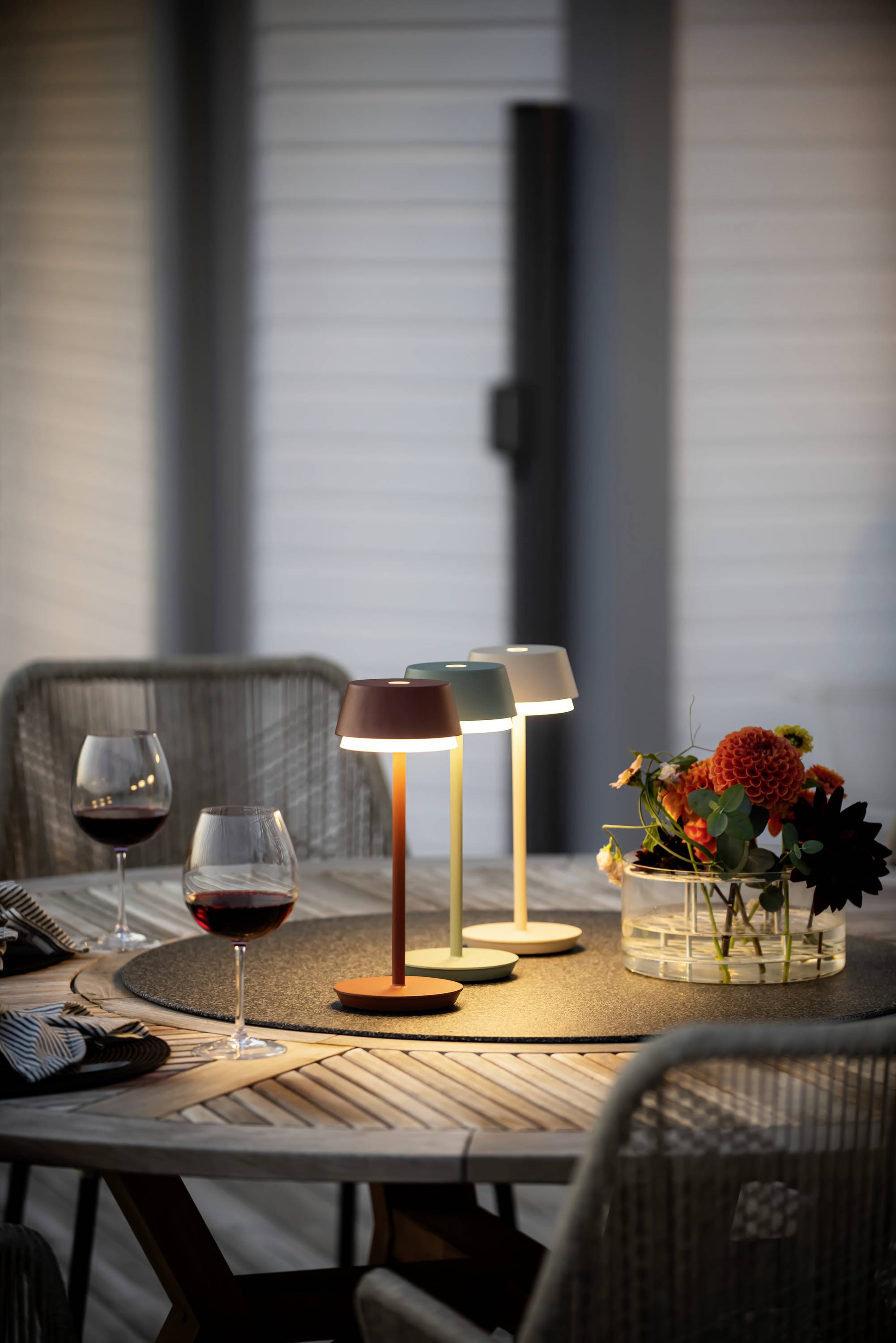 A table with three glowing, contemporary lamps, two wine glasses and a floral arrangement in a bowl at dusk.