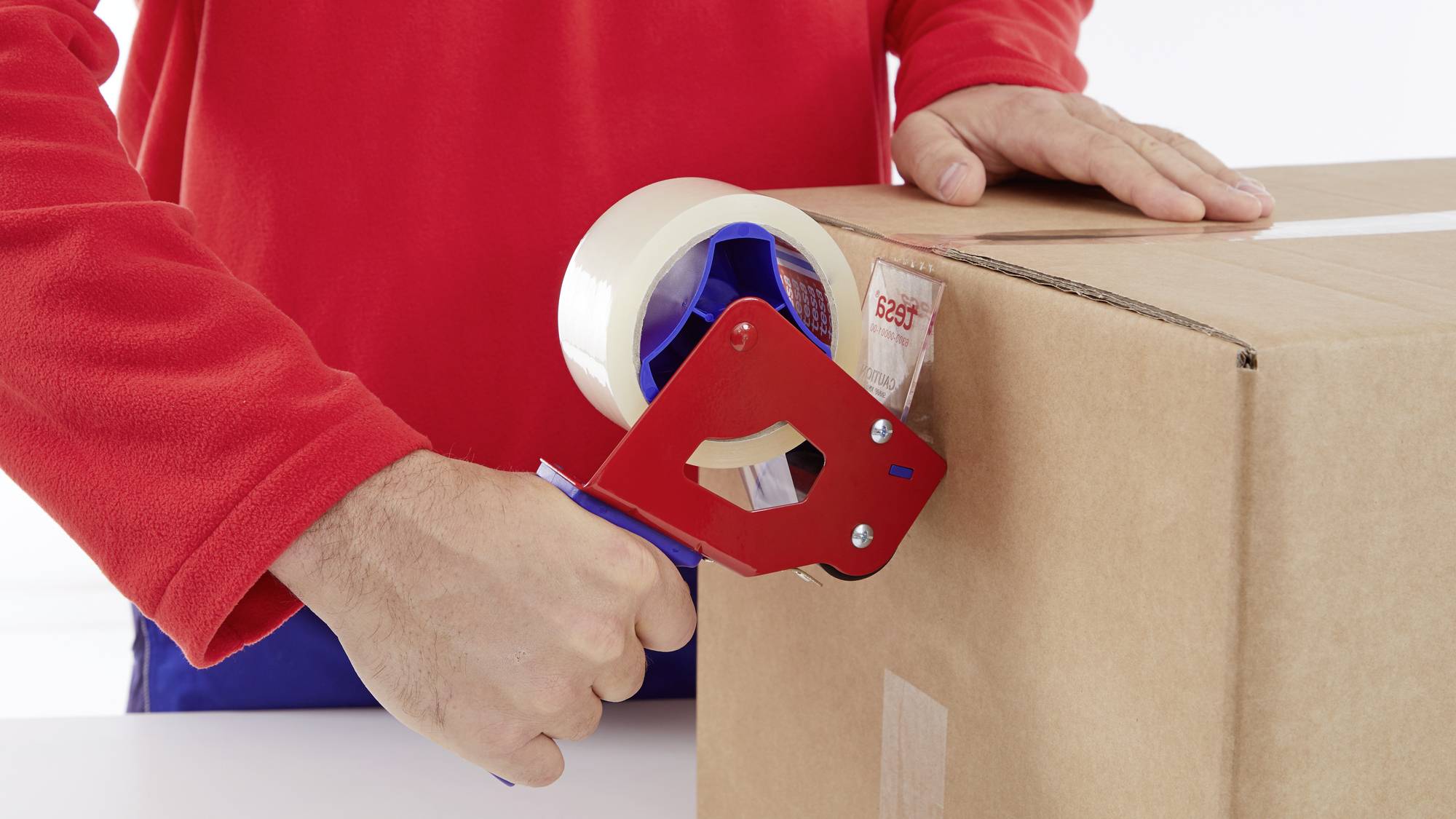 A person in a red top is sealing a cardboard box with a packing tape dispenser.
