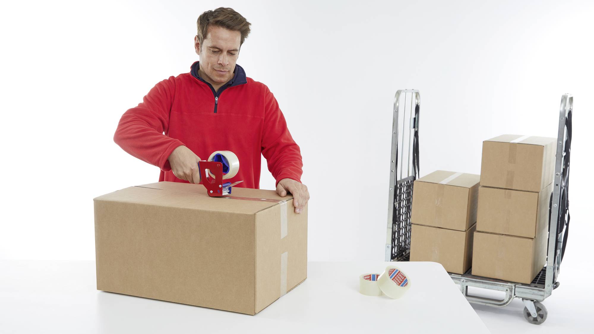 A man in a red jumper is sealing a cardboard box. In the background, there is a trolley with stacked boxes.