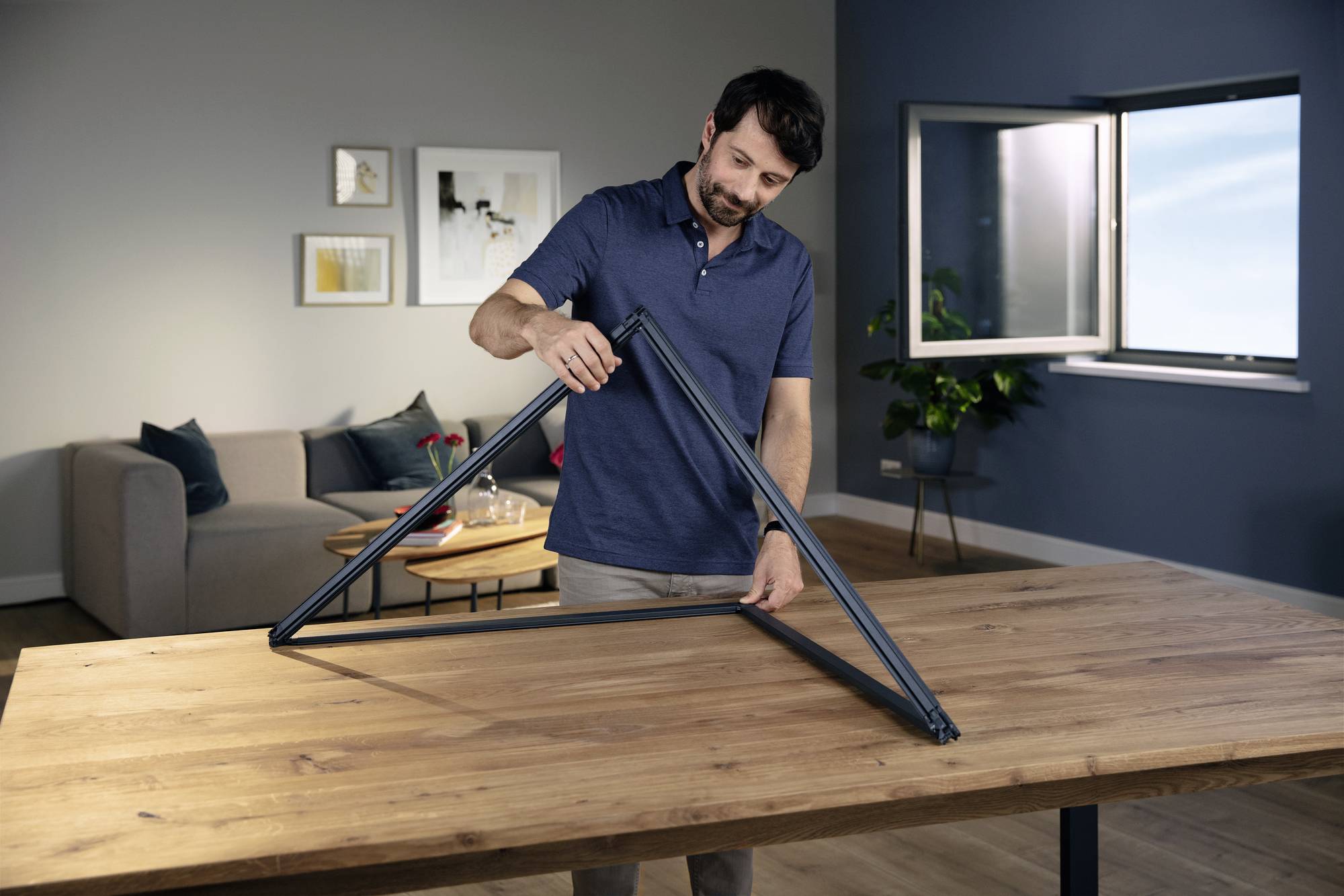 A man is assembling a metal frame on a table in a modernly furnished living room. A window is open.