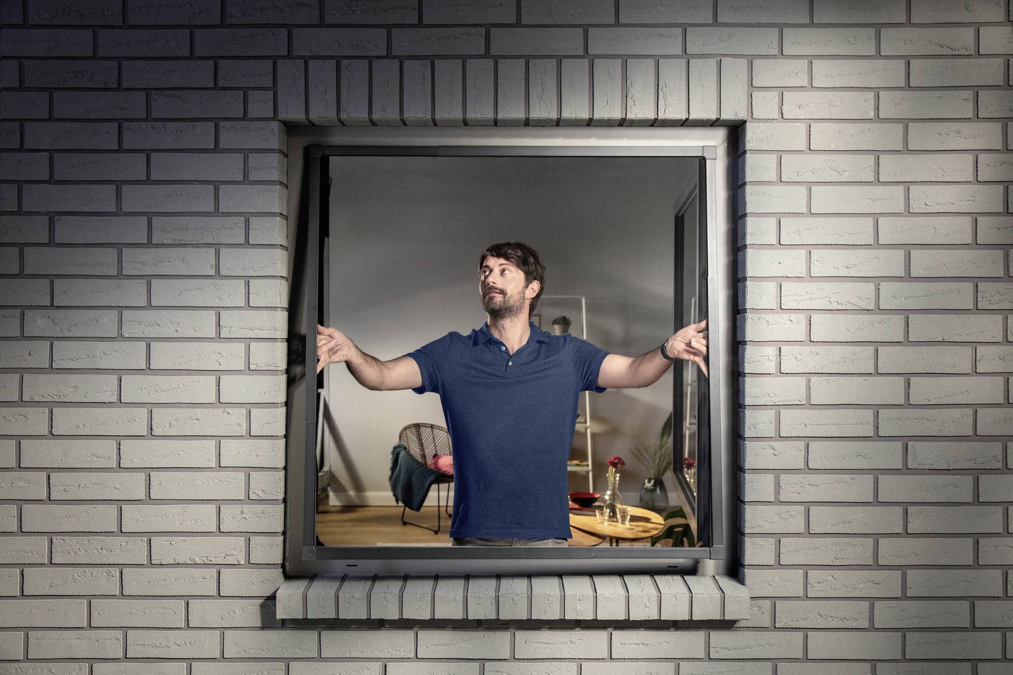 A man stands in an open window, spreading his arms and looking outside. Behind him is a modernly furnished room.