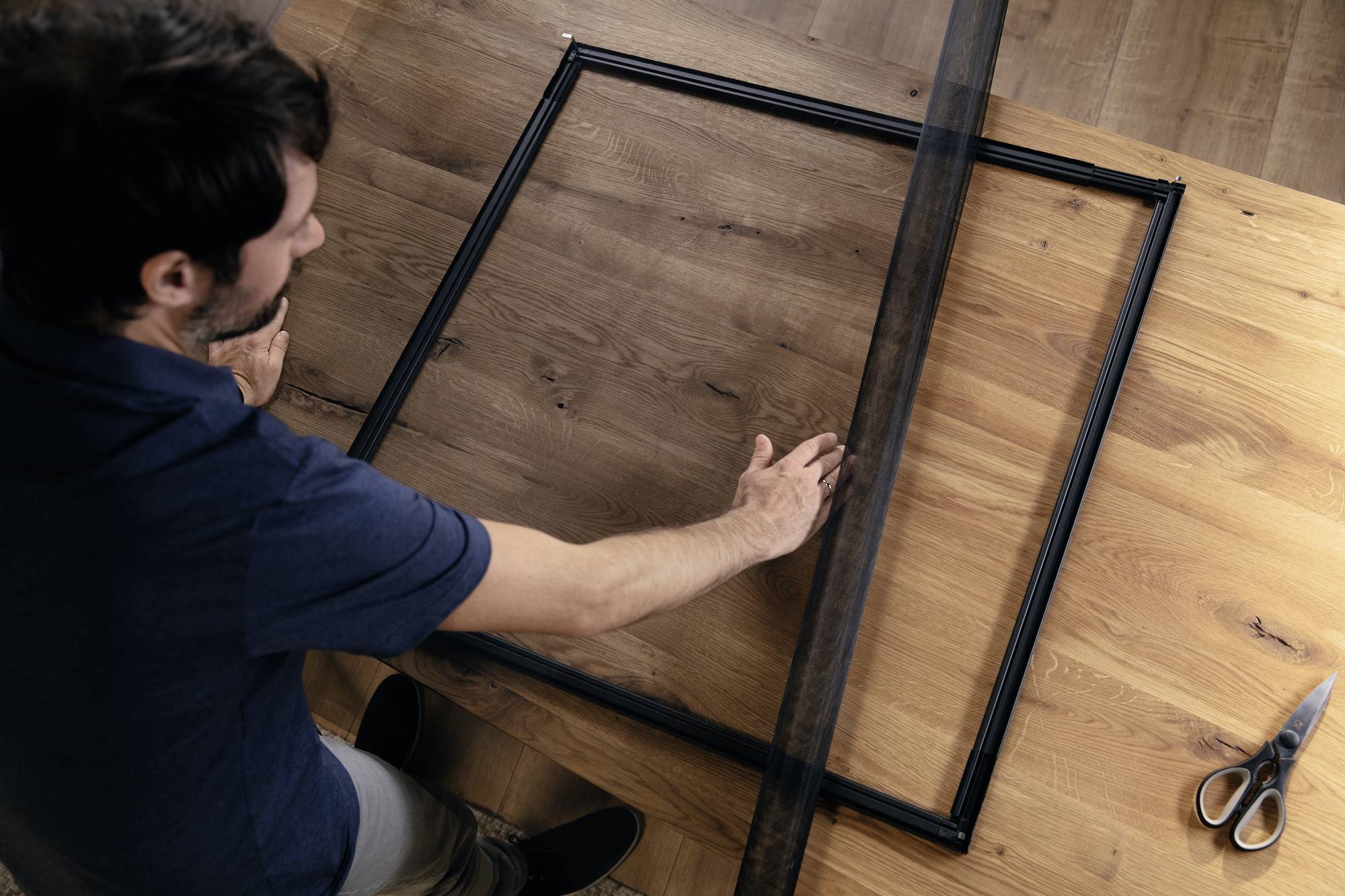 A man is working on a wooden frame on a wooden table, placing a piece of mesh fabric over it. A pair of scissors lies nearby.