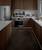 Modern kitchen with wooden cabinets, white worktop, stainless steel appliances and floor lighting. To the left, a fridge stands next to a cooker.