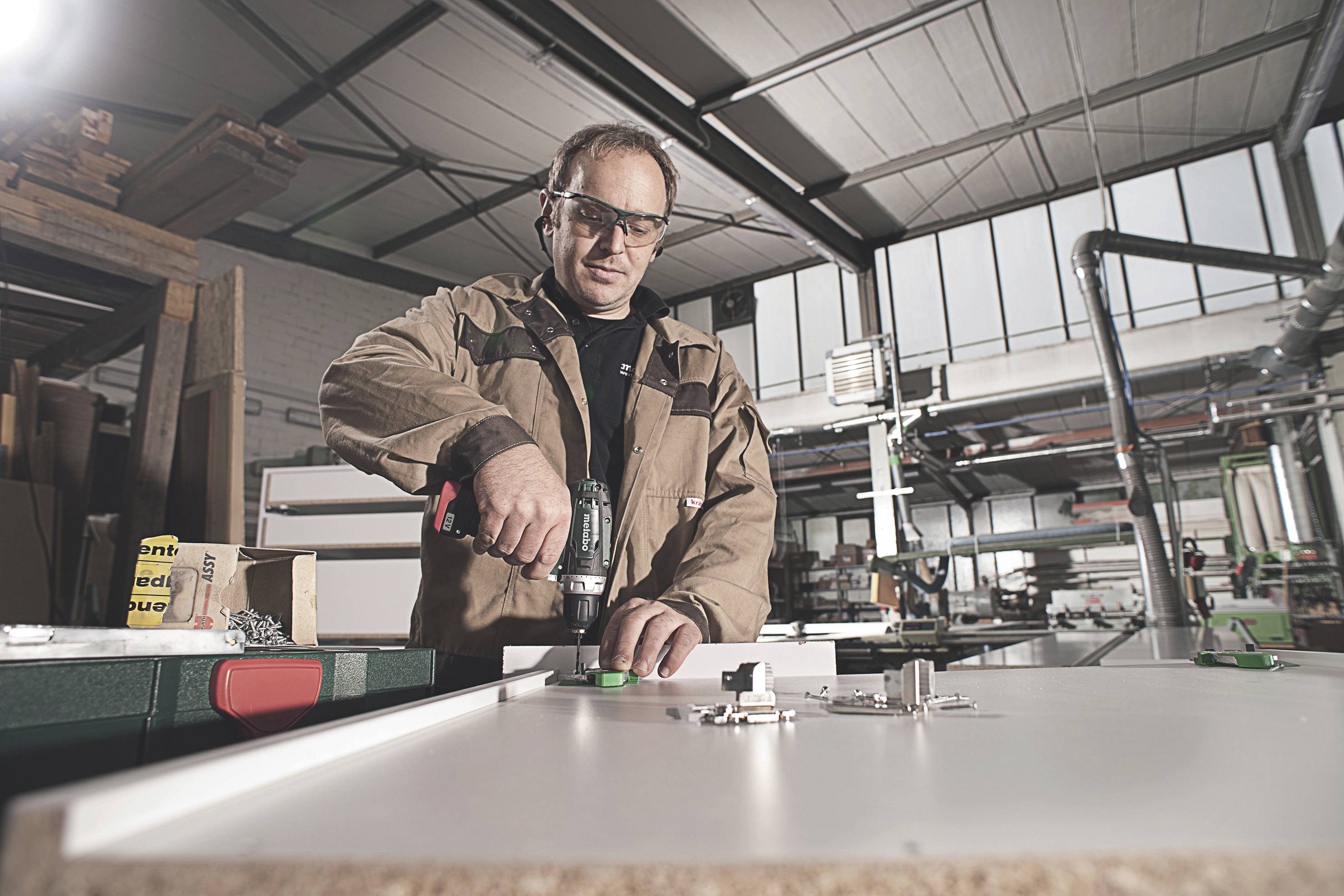 A person in work attire is drilling a hole into a white board with a cordless drill in a workshop. Tools are visible in the background.