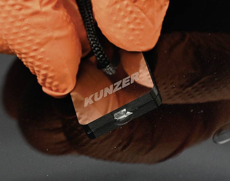 'Oranges Handschuh holds a small square mirror with the engraving 'KUNZER' near a dark surface.'