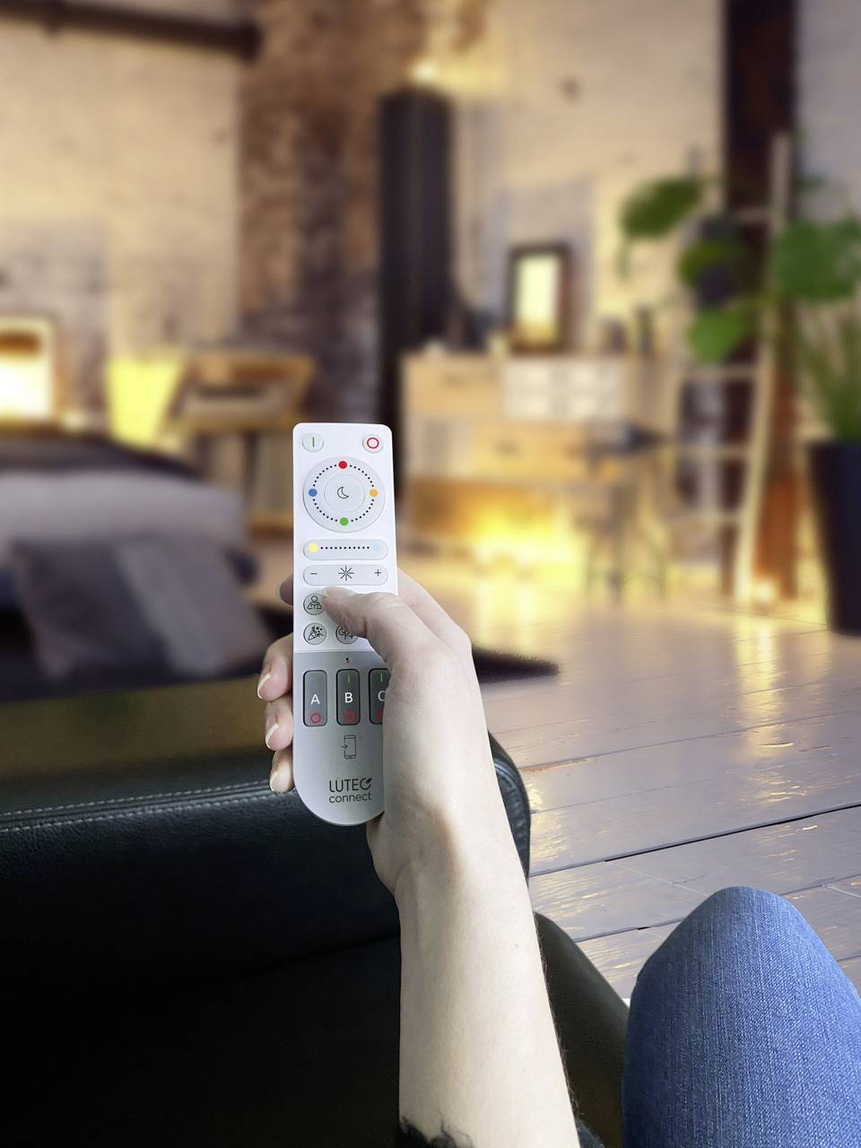 A person is holding a remote control in a cosy living room. A bed and plants are visible in the background.