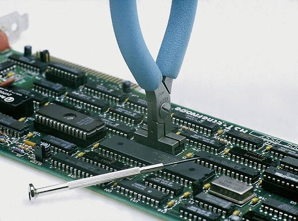 A pair of pliers is holding a component on a circuit board, with a small screwdriver lying beside it.