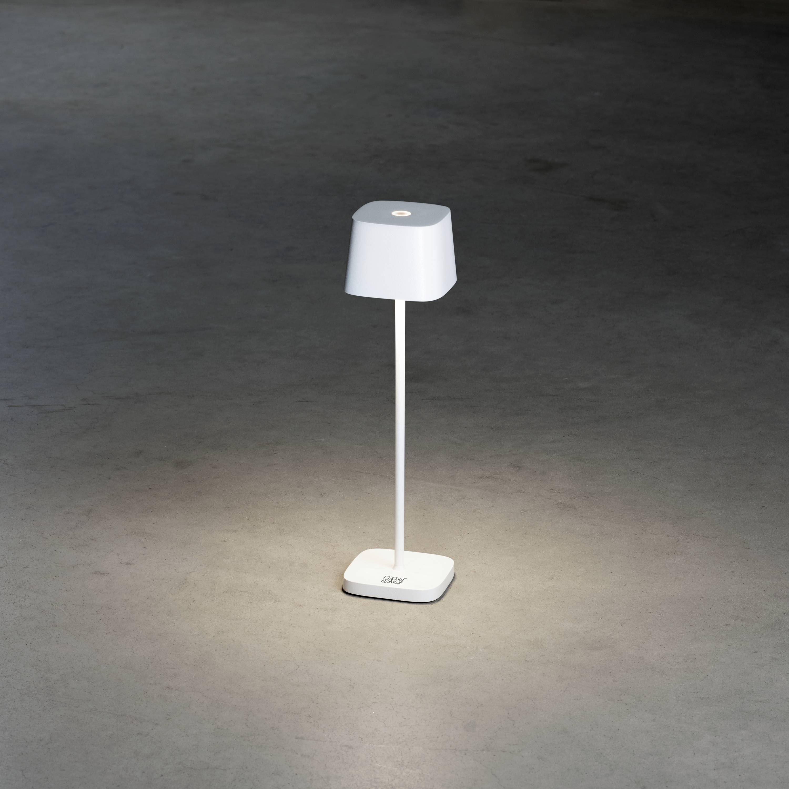 White table lamp with a slender neck and wide shade, illuminated against a dark background. Modern, minimalist design.