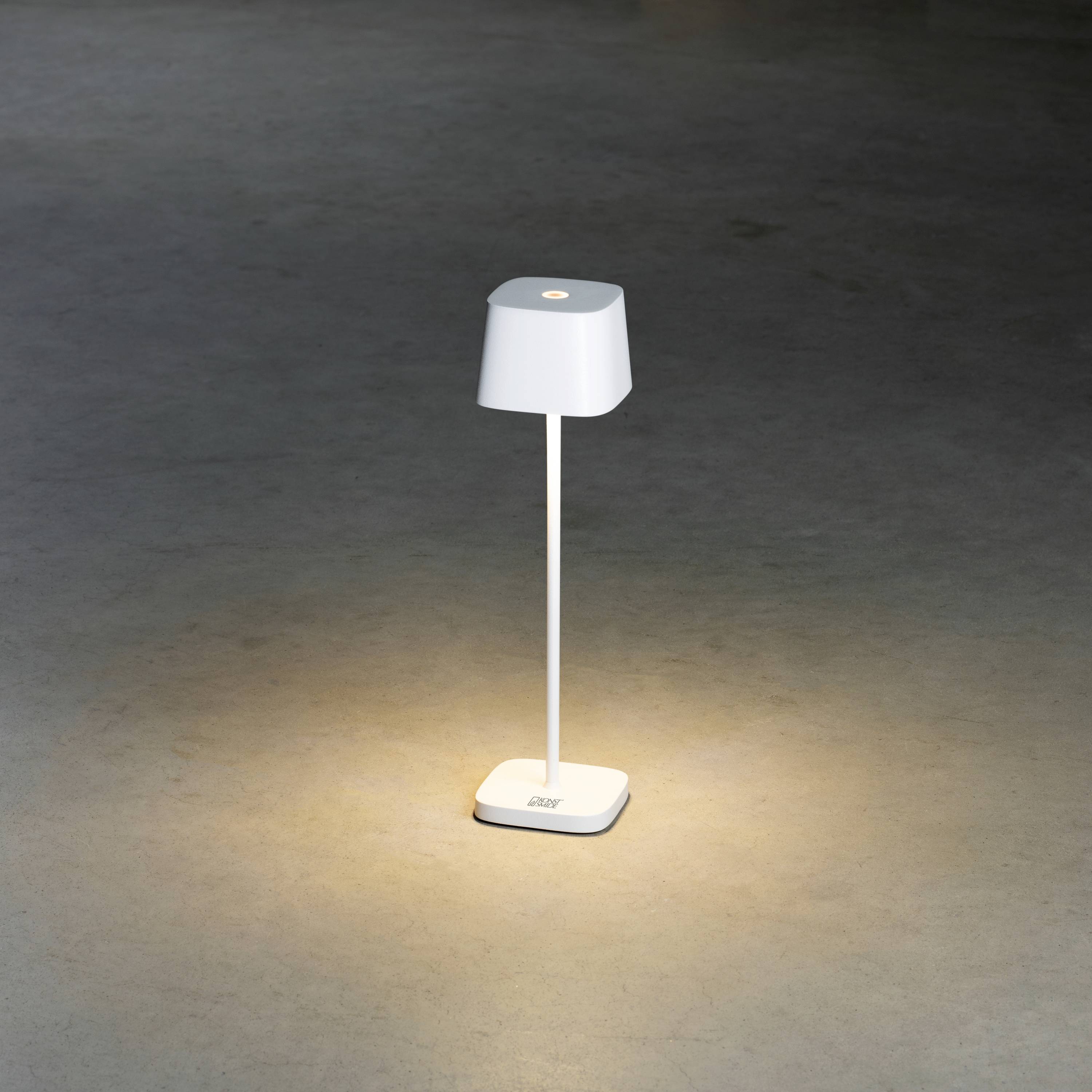 White, modern table lamp on grey floor; casts soft, warm light in a circular area around itself.