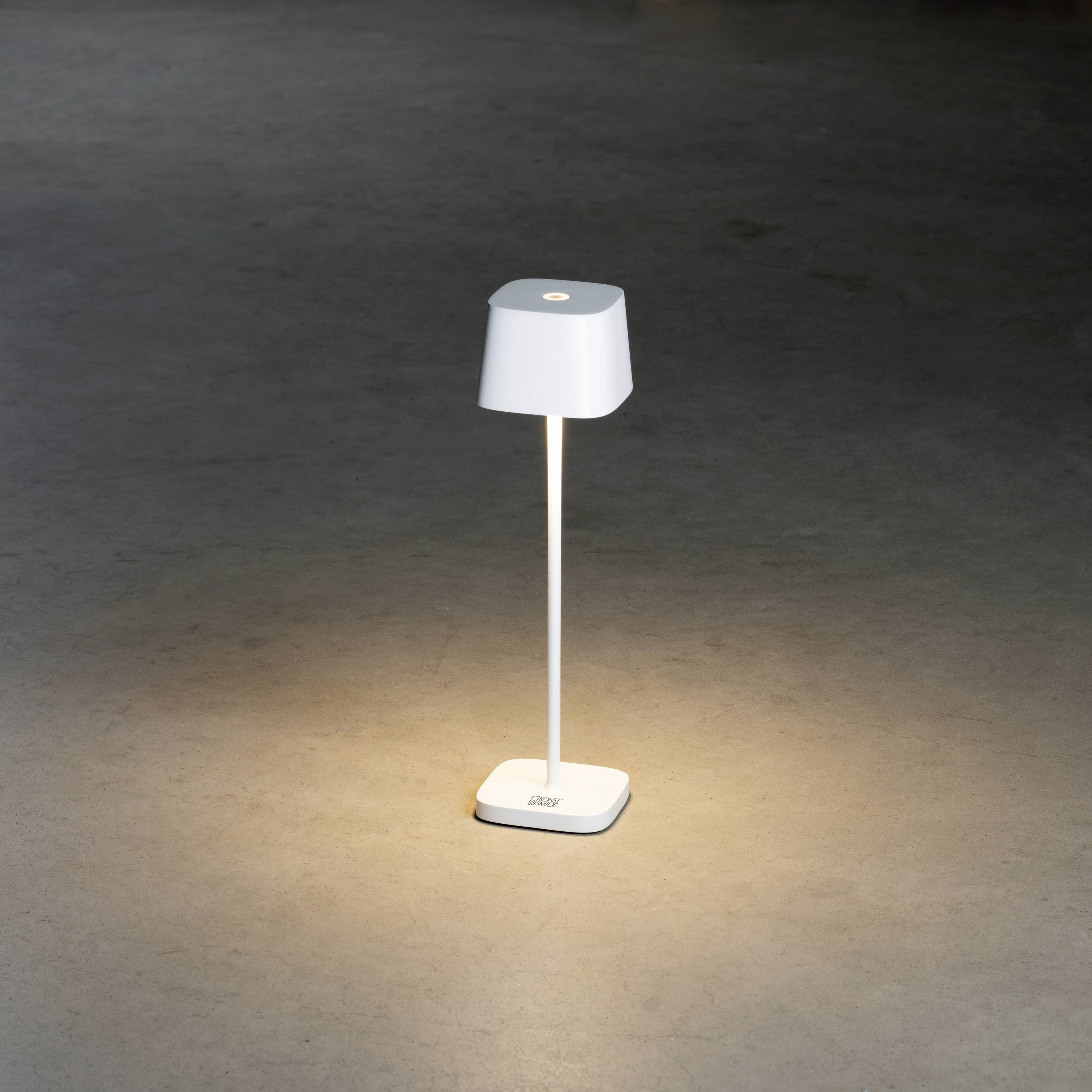 A simple, white table lamp with a round shade illuminates a dark room. The lamp sits on a light rectangular base.