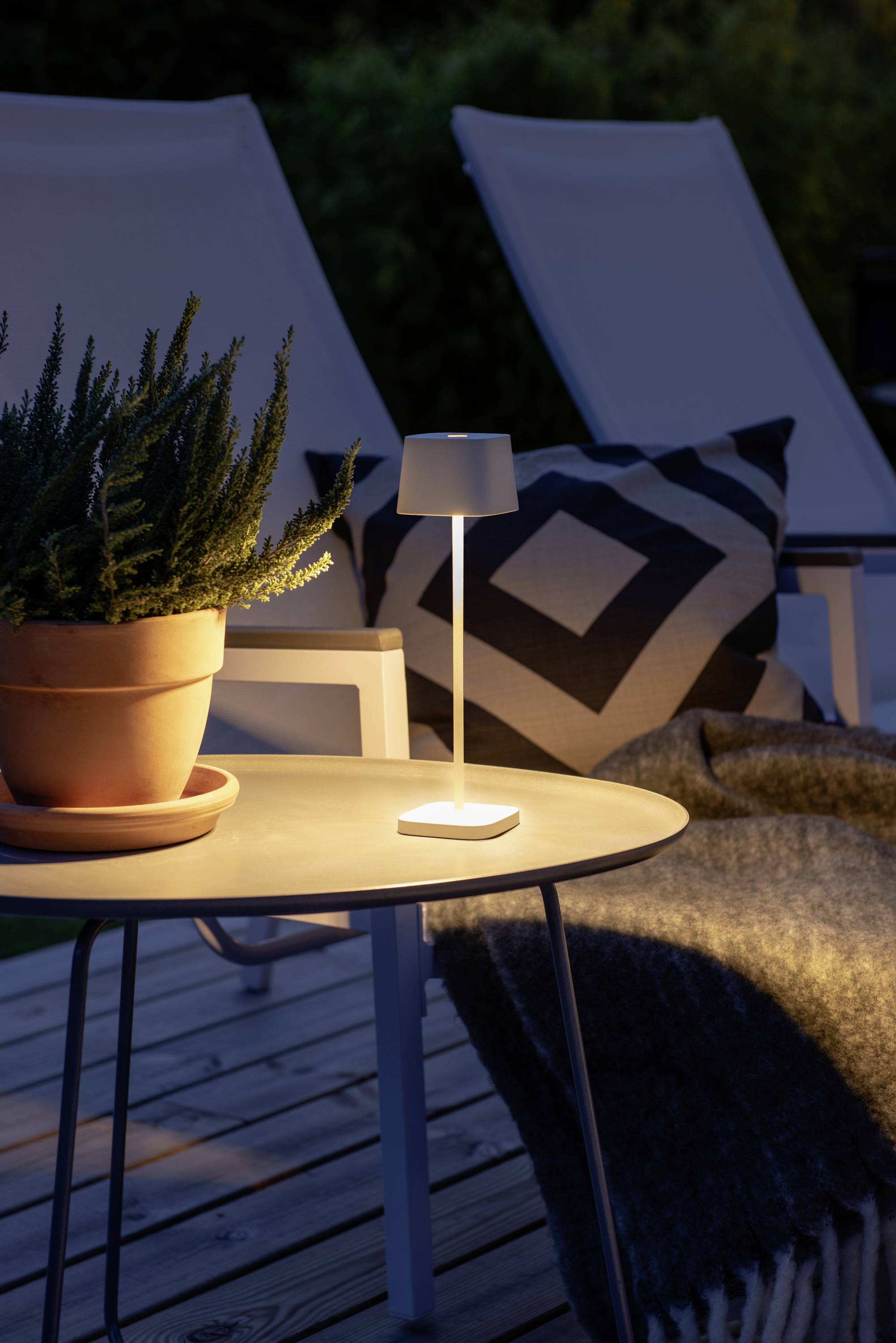 An outdoor table with a lamp, a plant pot, and a chair in the background. Cosy atmosphere at dusk.