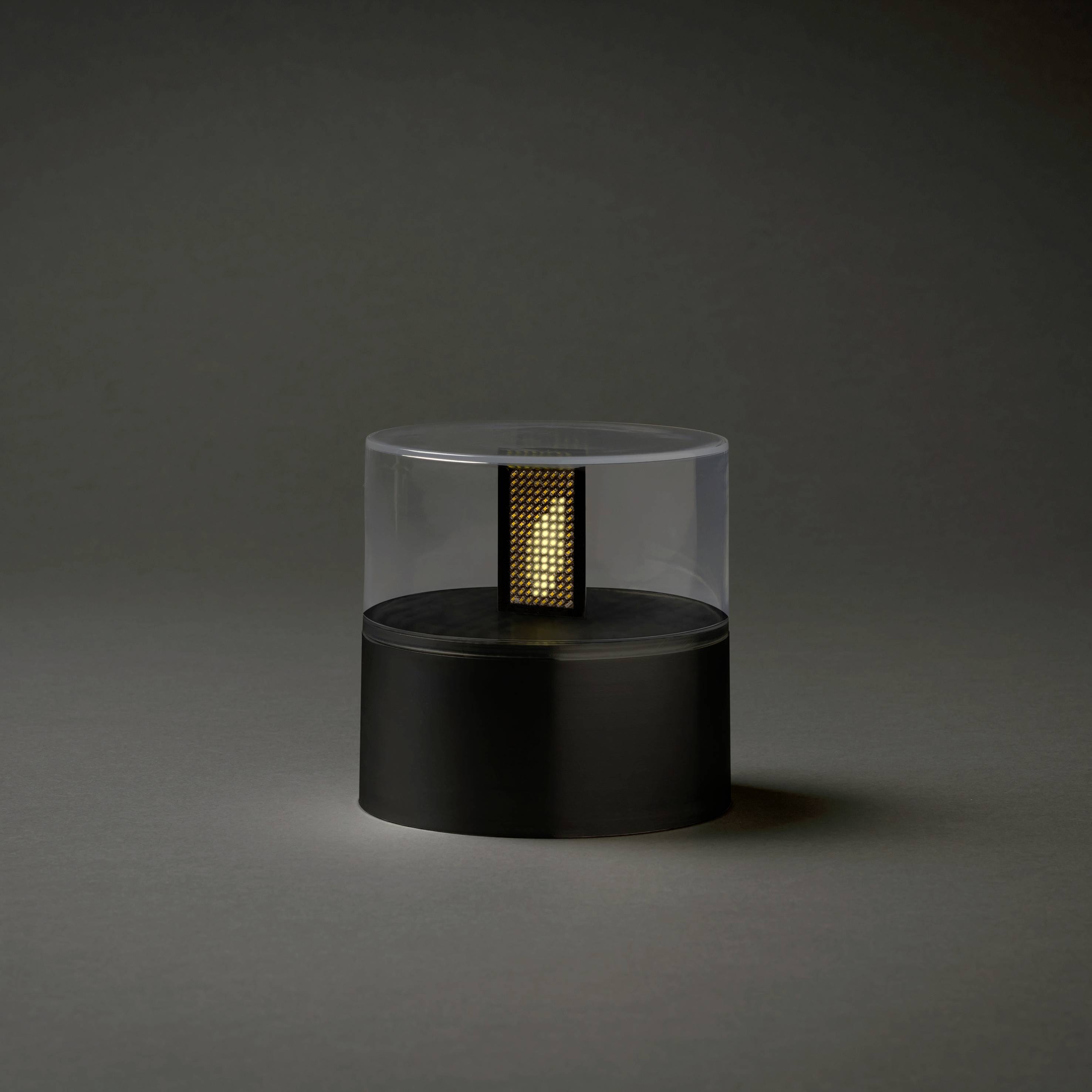 Small, round, black lamp with a clear cylindrical casing and inner golden grid, against a dark background.