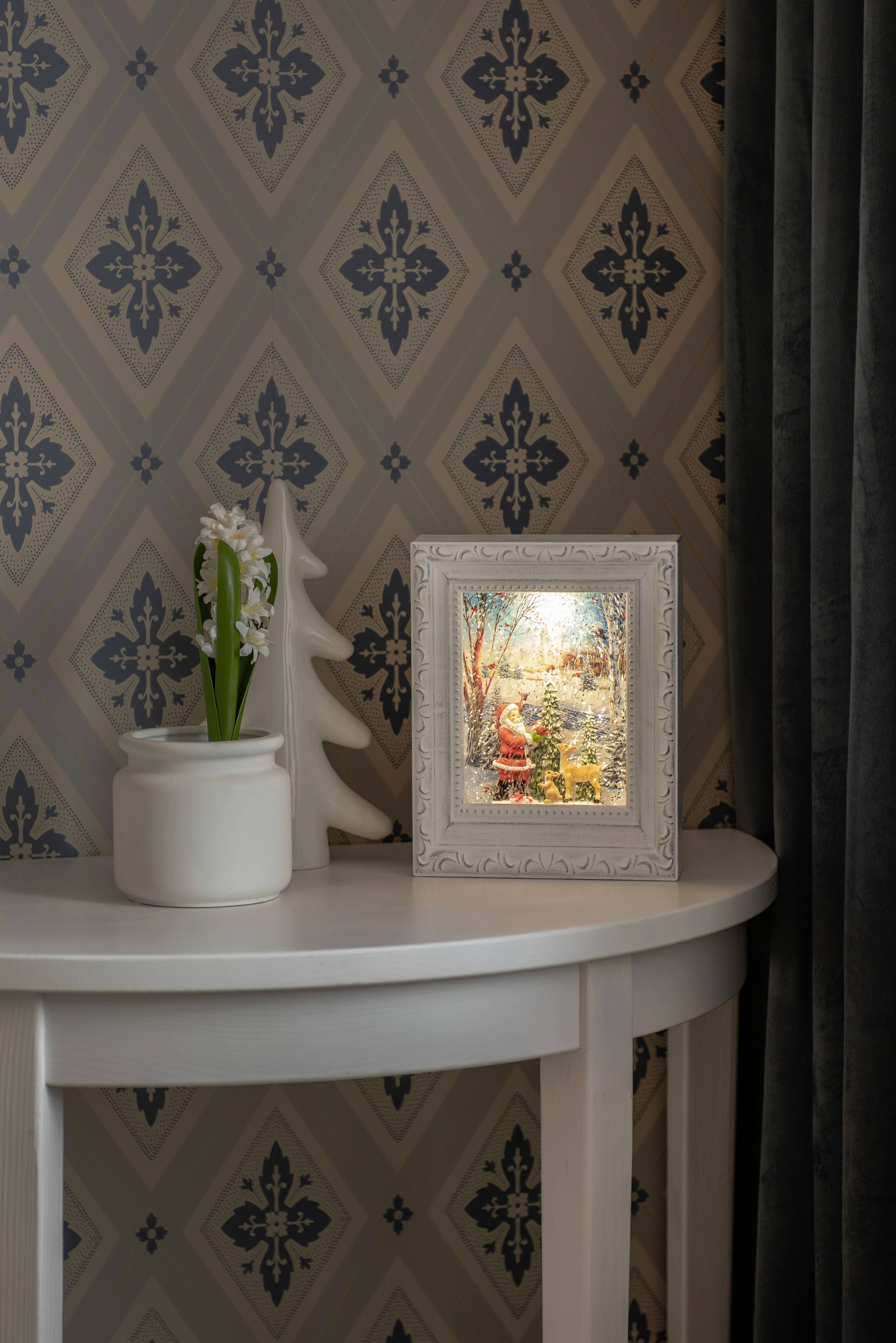 A small, decorative table stands against a patterned wallpapered wall, with a white vase of flowers and a framed photograph in the foreground.