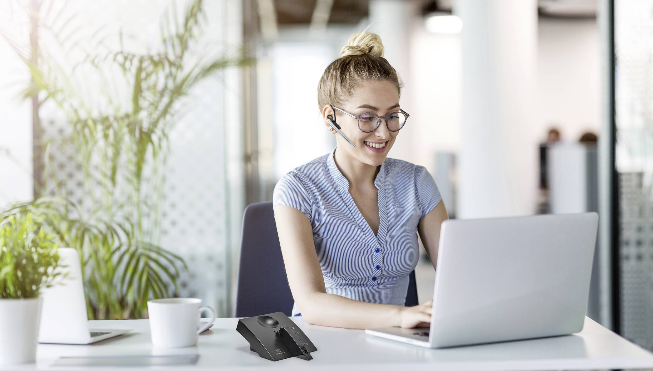 A woman in the office wearing a headset is working on a laptop, with a telephone on the desk.