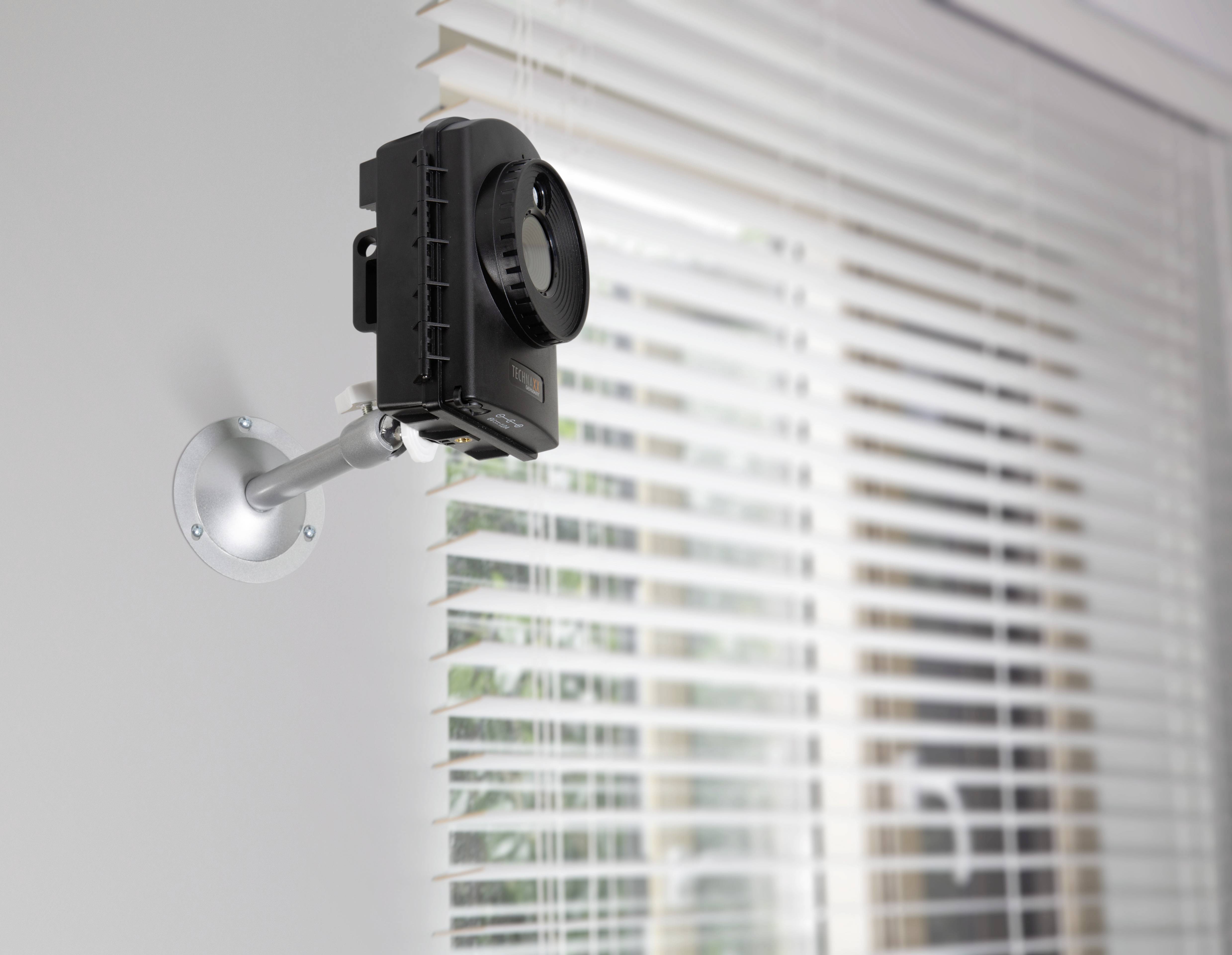 A black camera is mounted on the wall next to a window with venetian blinds.