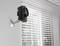 A black camera is mounted on the wall next to a window with venetian blinds.