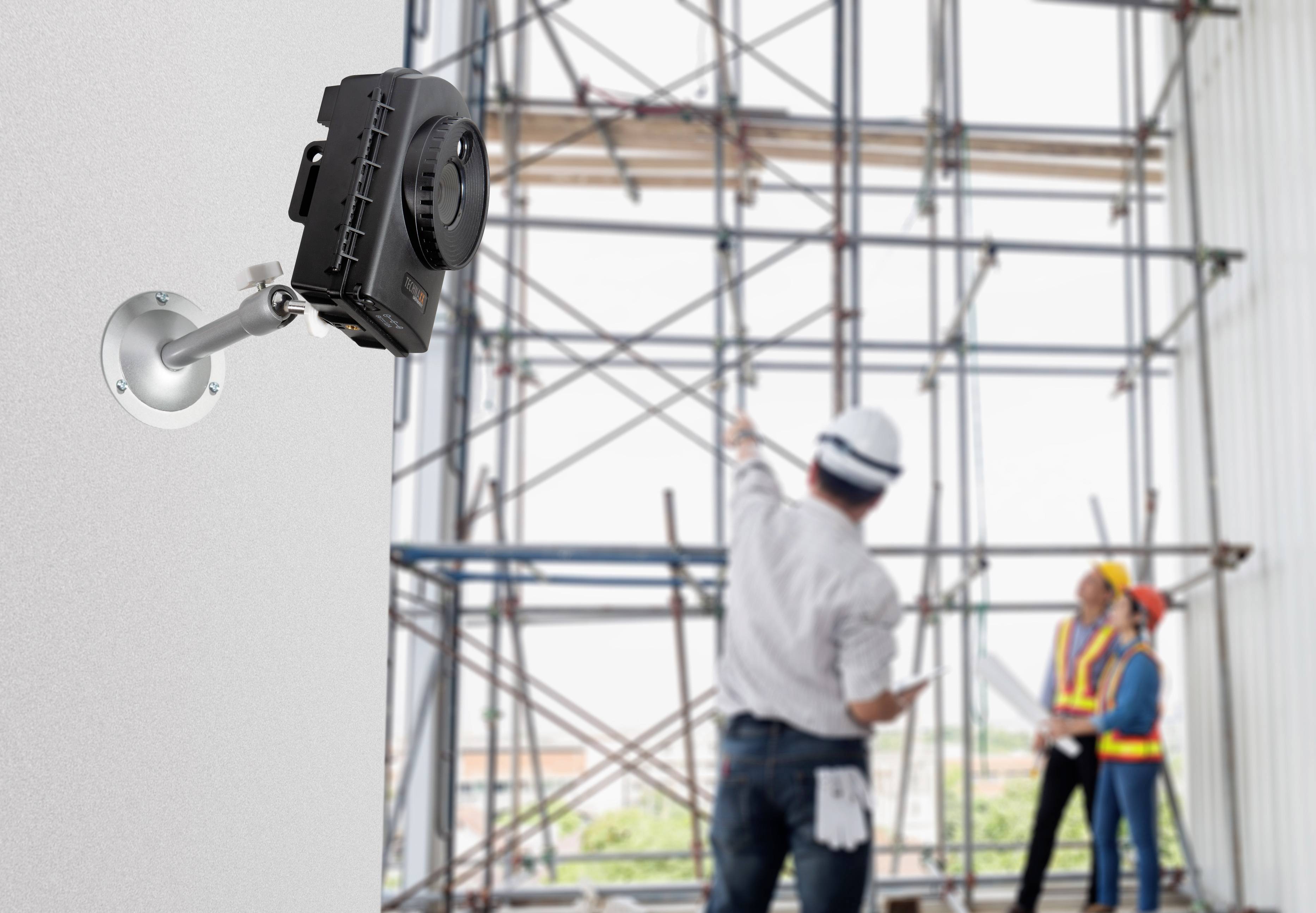 A surveillance camera is mounted inside, with construction workers in conversation in front of a scaffolding in the background.