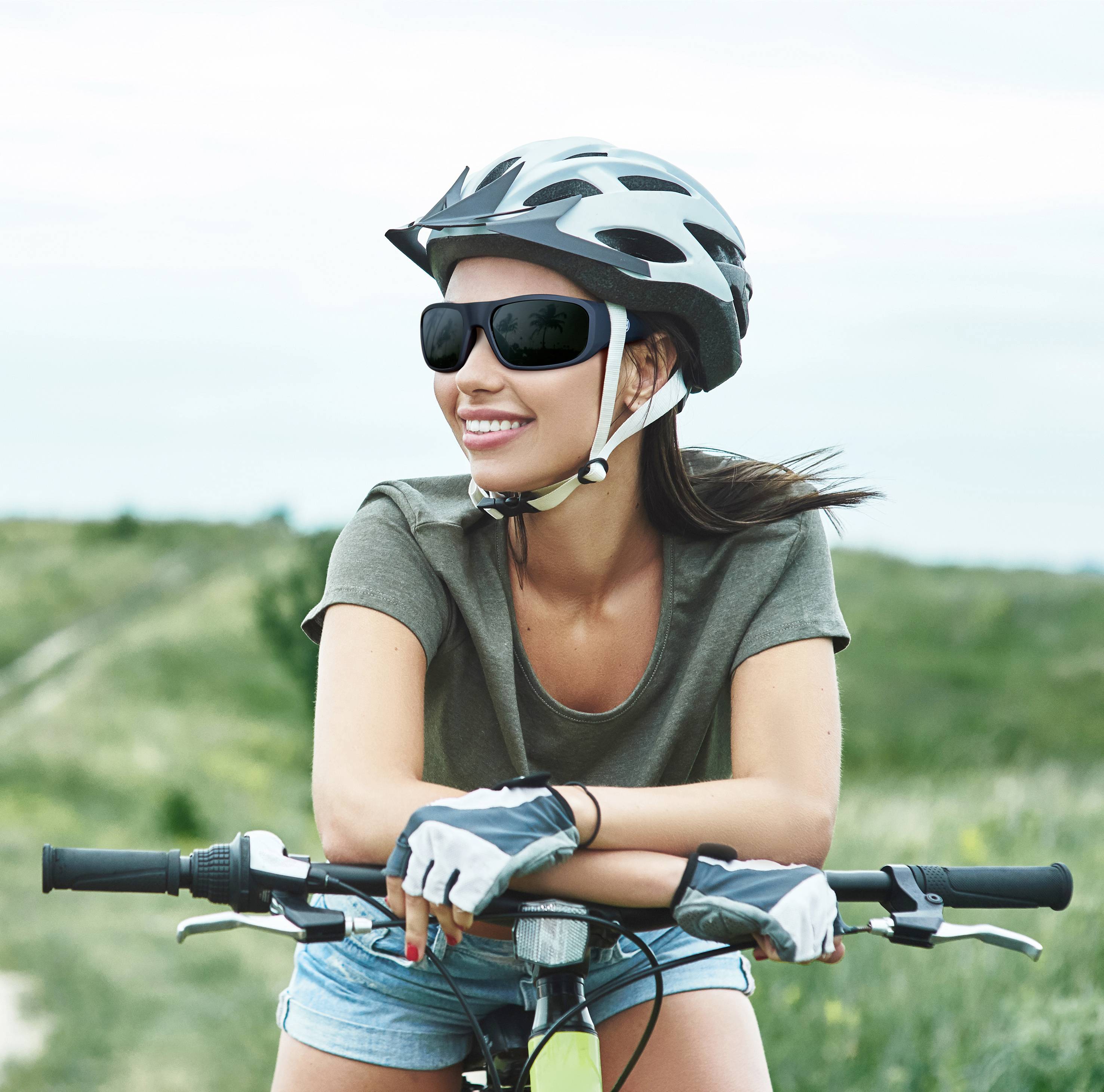 A person wearing a helmet and sunglasses is cycling with a smile through a green, rural landscape.