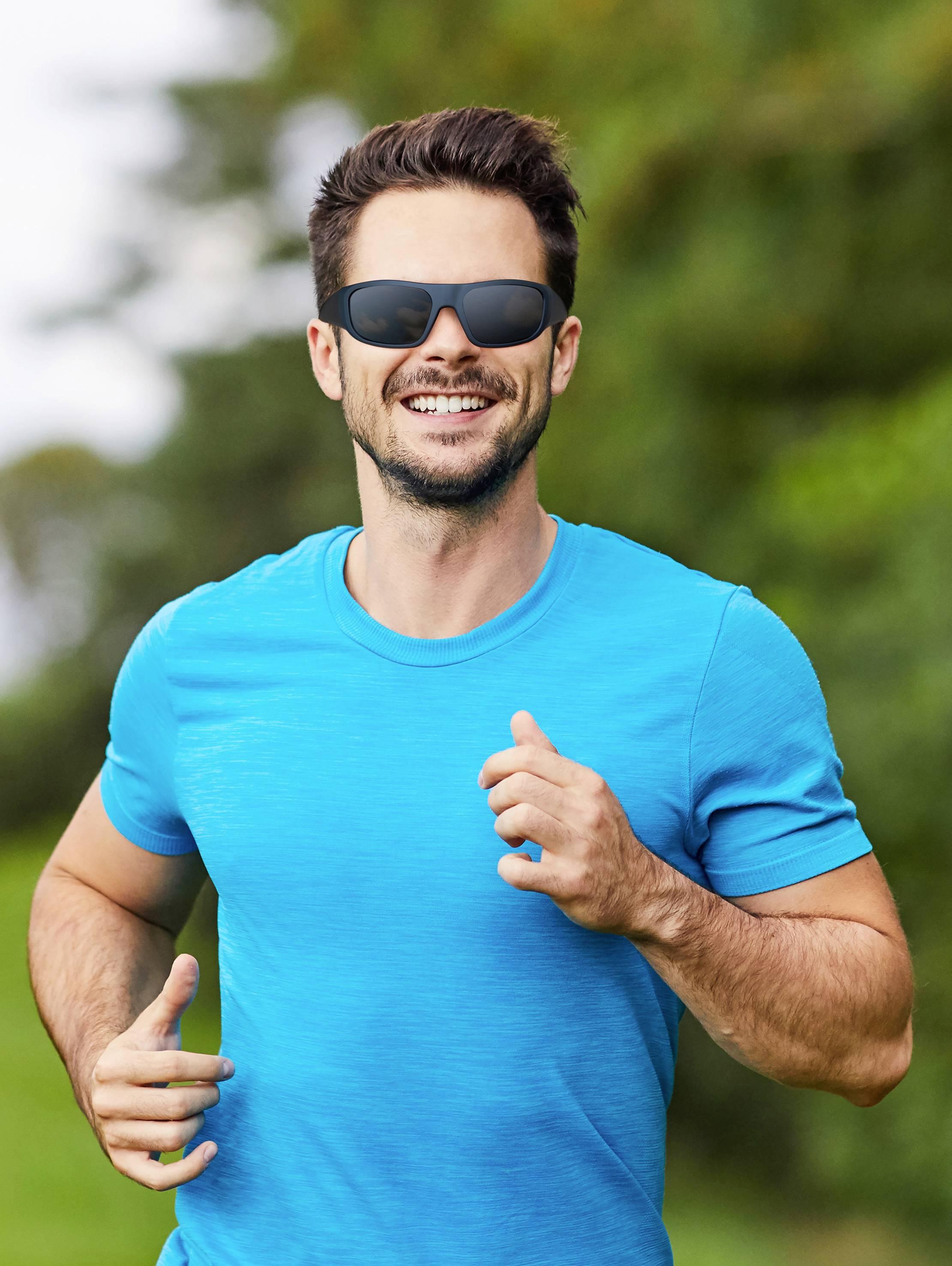 A man in a blue t-shirt is jogging outside on a sunny day, wearing sunglasses and smiling. Trees can be seen in the background.