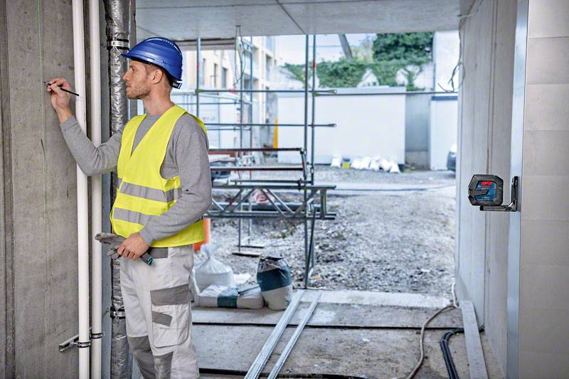 A construction worker wearing a blue hard hat and yellow high-visibility vest is making notes on a wall. Scaffolding and building materials are visible in the background.