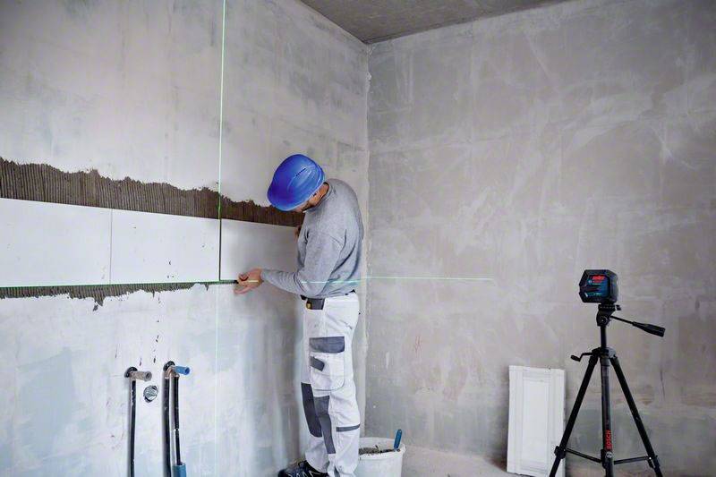 A tradesman wearing a hard hat is measuring a wall with a laser tool to install pipes. A tripod with a laser device stands nearby.