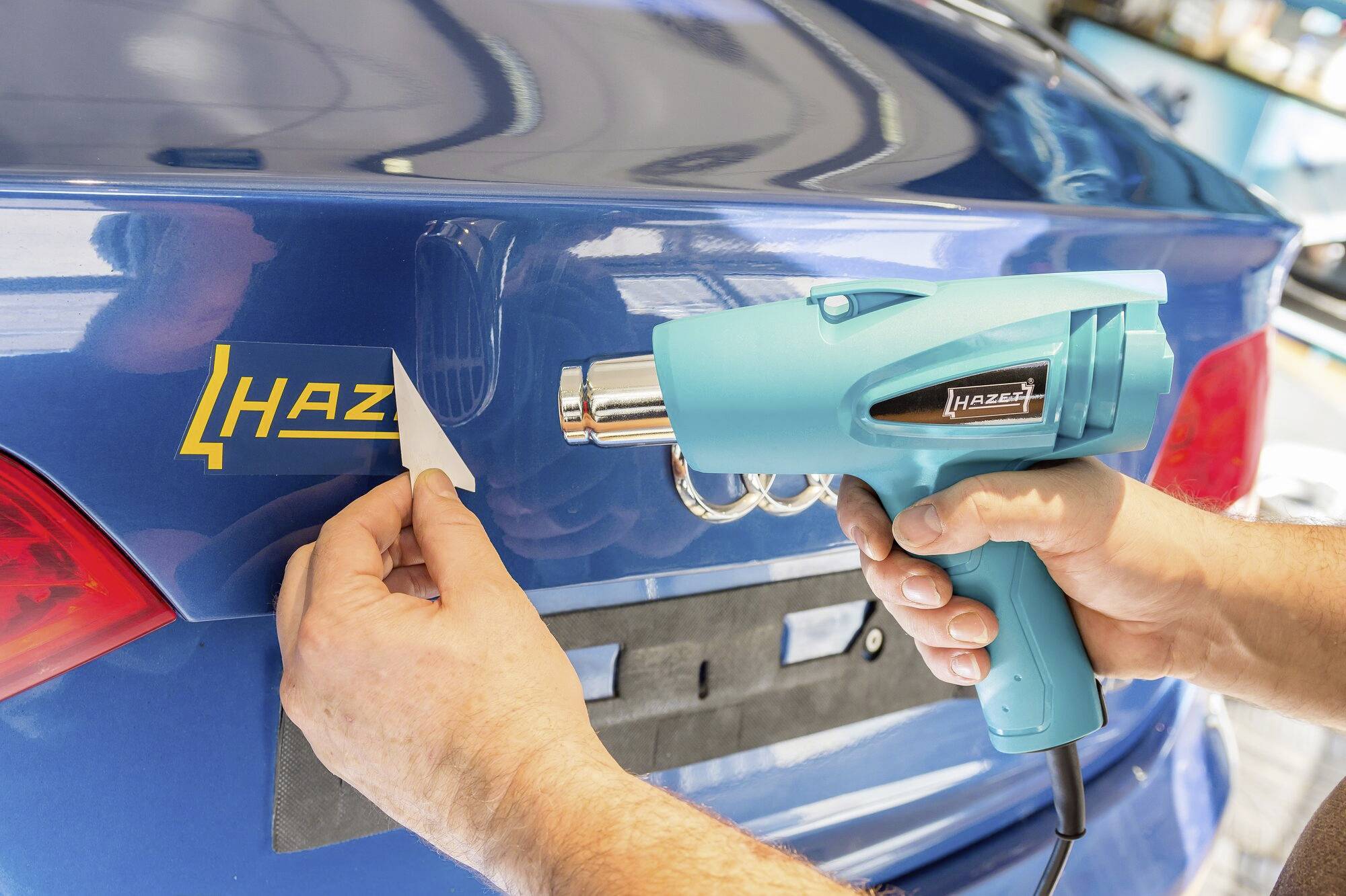 A person is removing a sticker from a blue car using a heat gun.