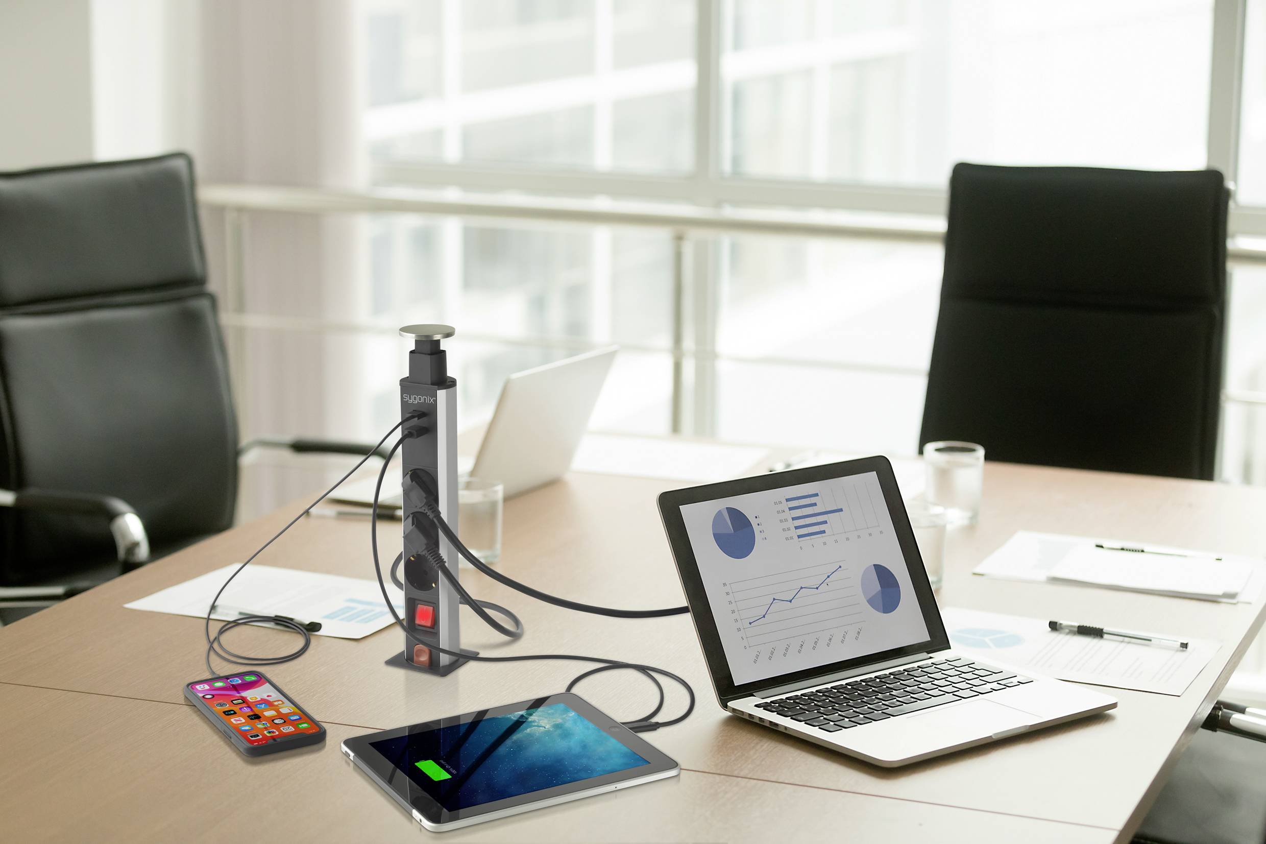 A conference table with a laptop, tablet and smartphone connected via a power strip. The laptop displays charts.