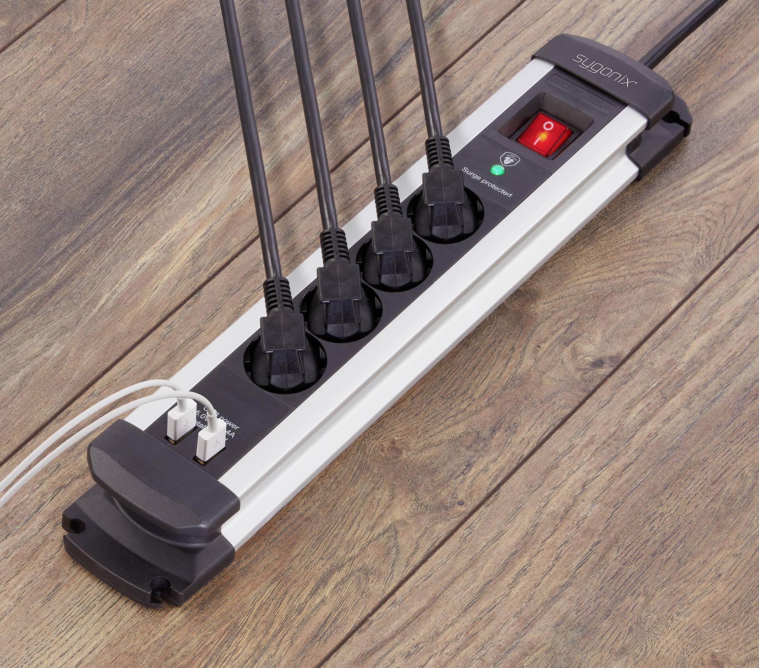 A black power strip with five devices plugged in lies on a wooden floor. A red switch is set to 'On'.
