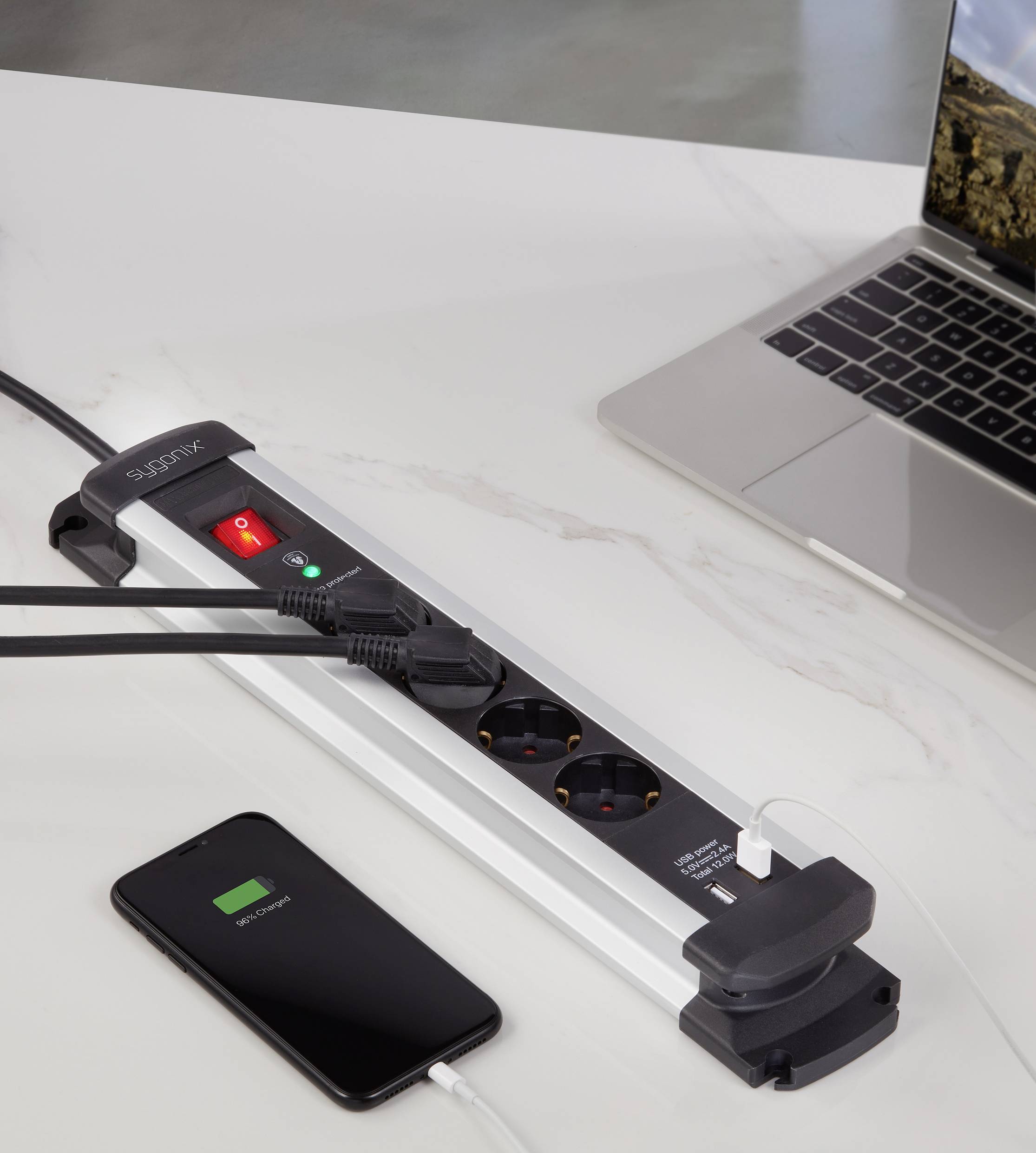 A multi-socket extension lead with its switch turned on is supplying power to a laptop and a charging smartphone on a table.