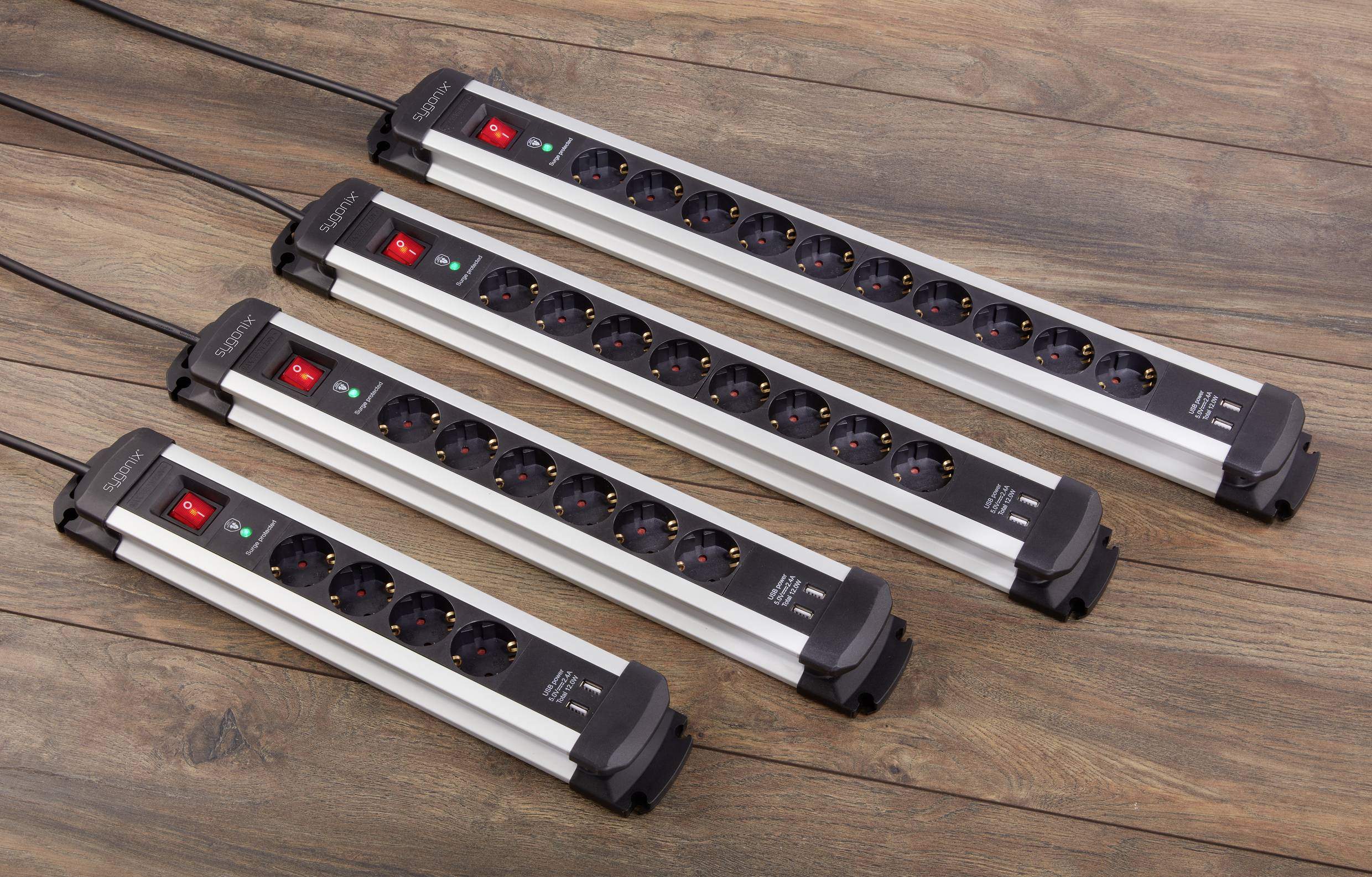 Four grey multi-socket extension leads with power switches and LEDs, lying on a wooden floor. Different lengths, suitable for multiple devices.