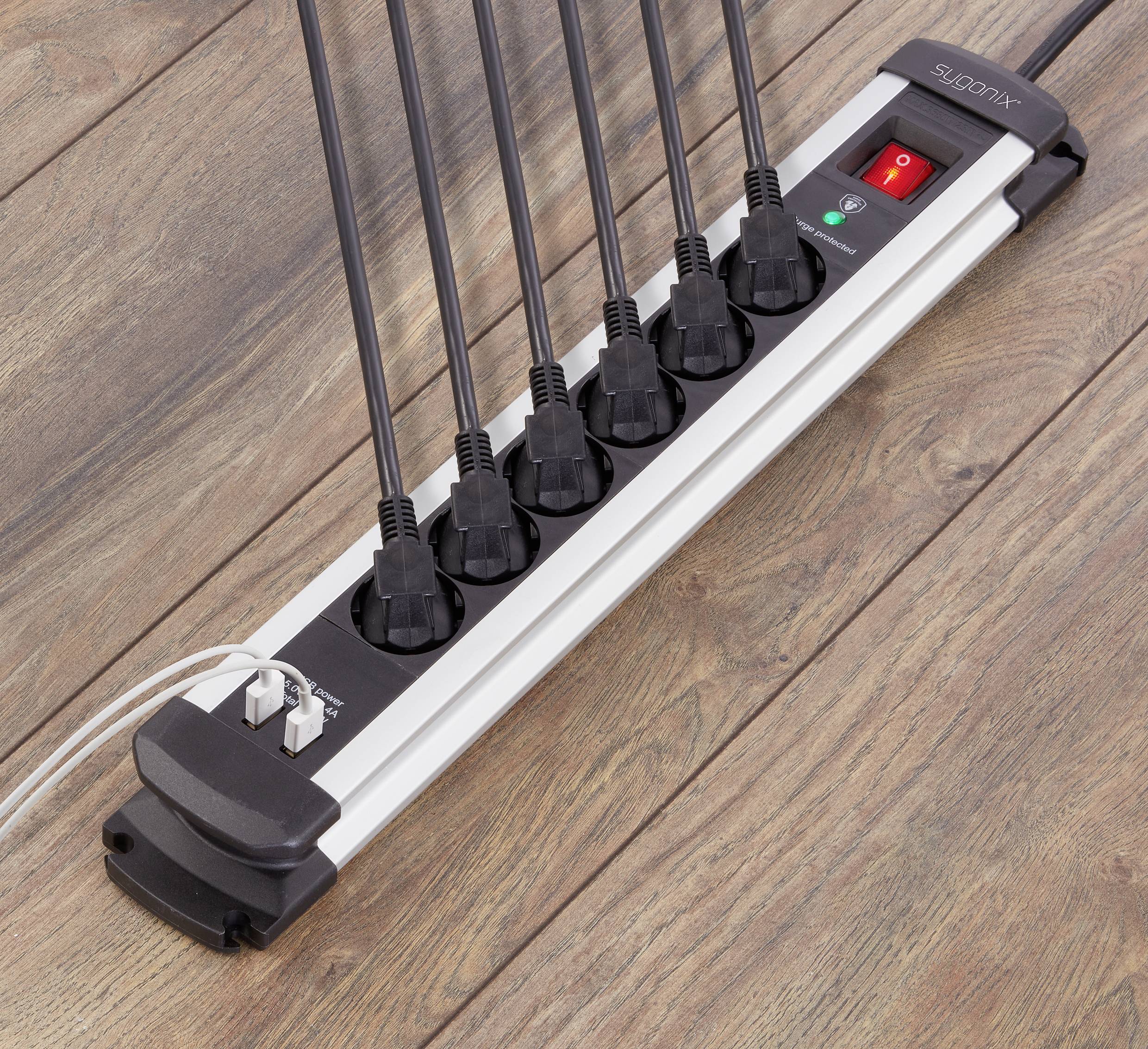 A multi-socket extension lead with seven sockets on a wooden floor. All socket outlets are occupied by black plugs. One switch is active.