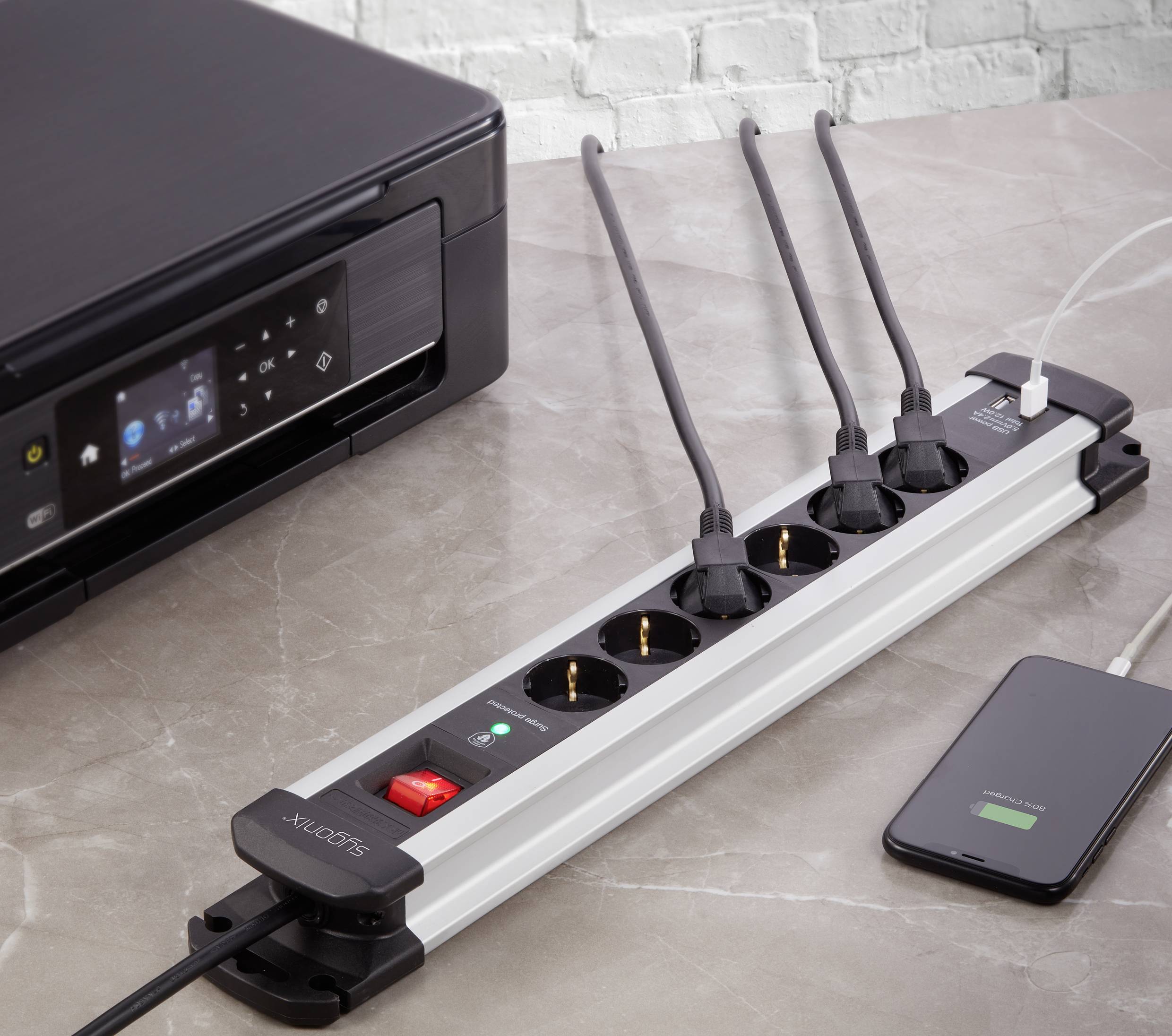 A table with a printer, a four-socket extension lead with four plugged-in power cables and a mobile phone charging.