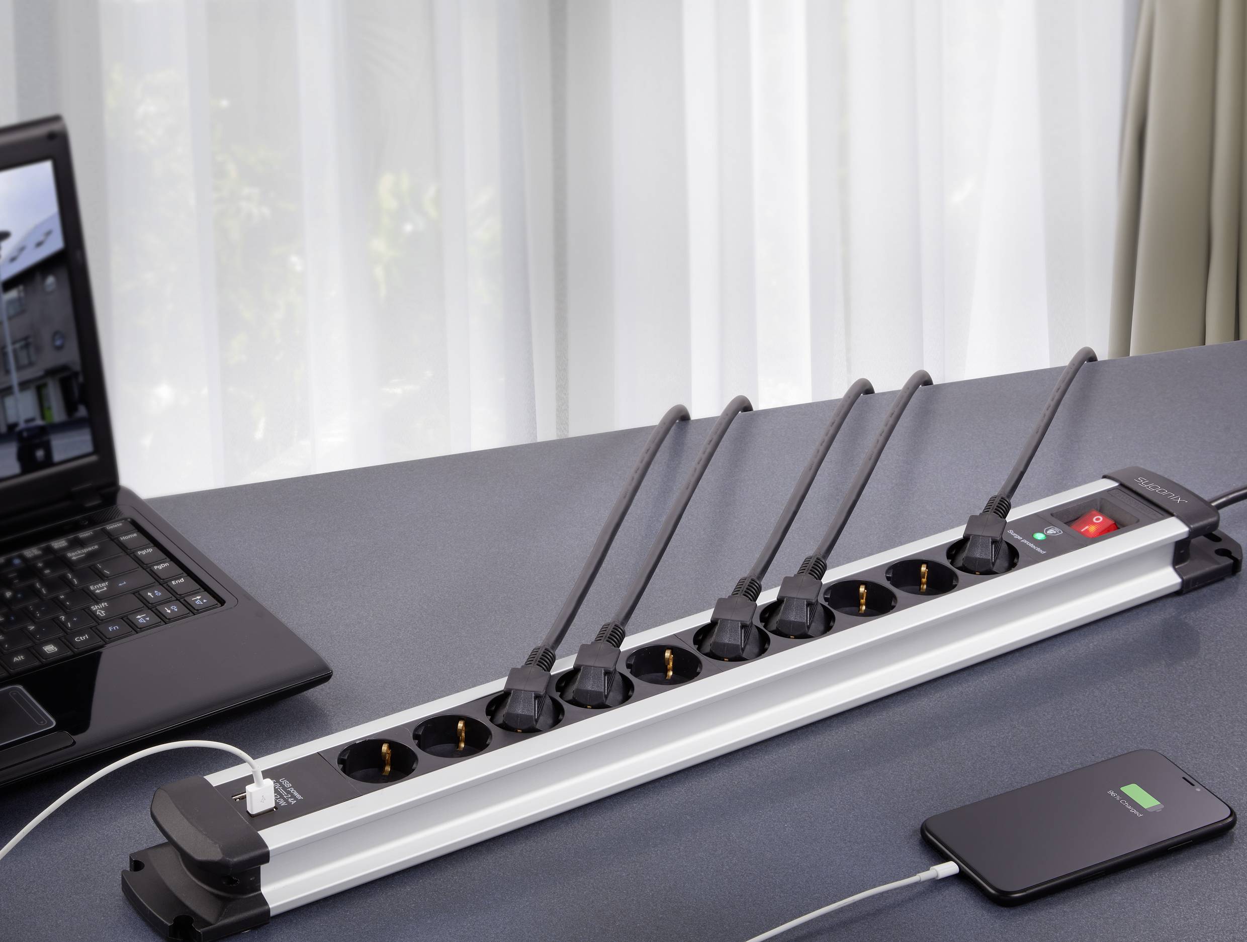 Multiple power socket on a table, with five devices connected. Laptop and smartphone are visible, smartphone is being charged.