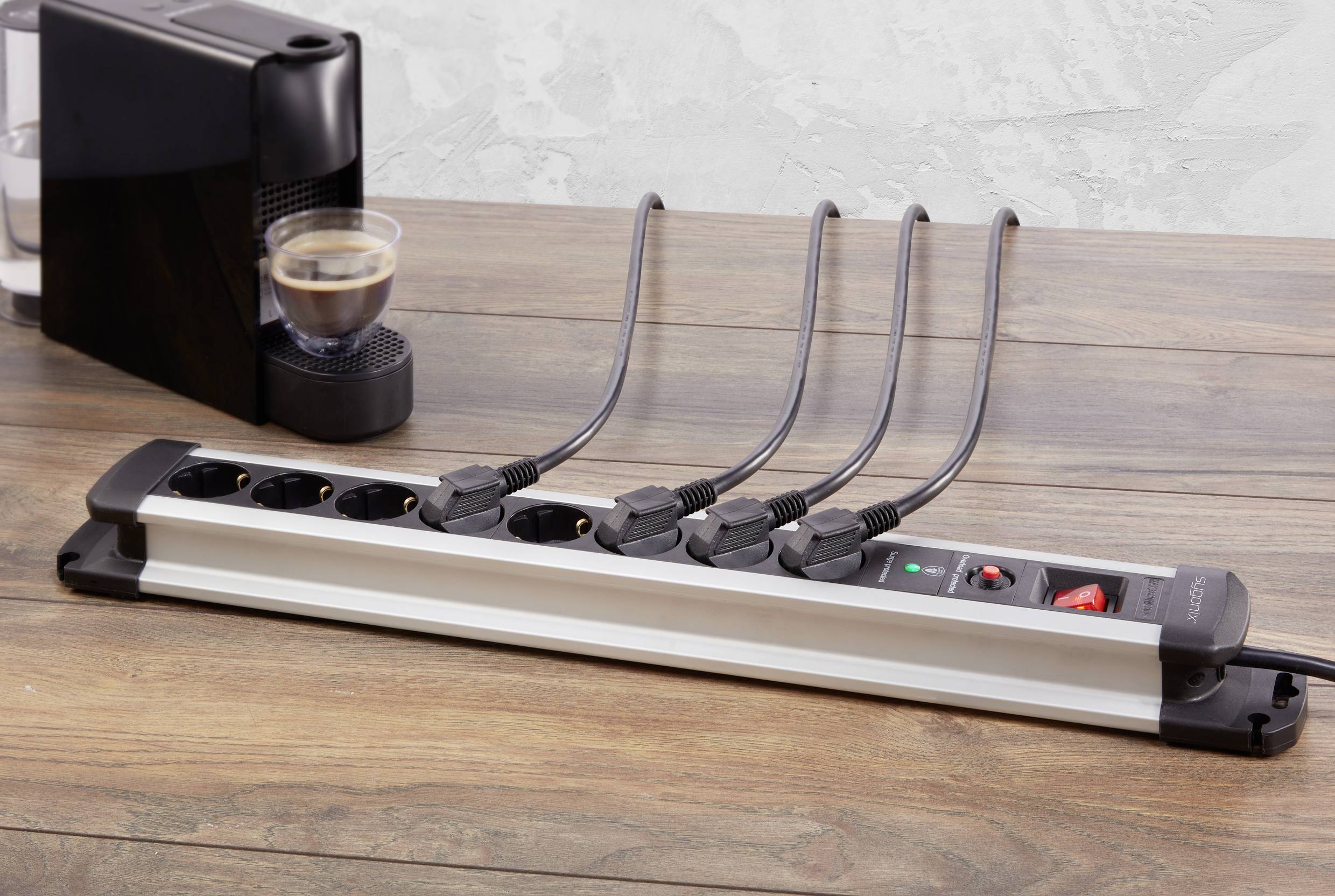 Multiple socket extension lead on a wooden table with six cables plugged in. In the background, a coffee machine with a cup stands.
