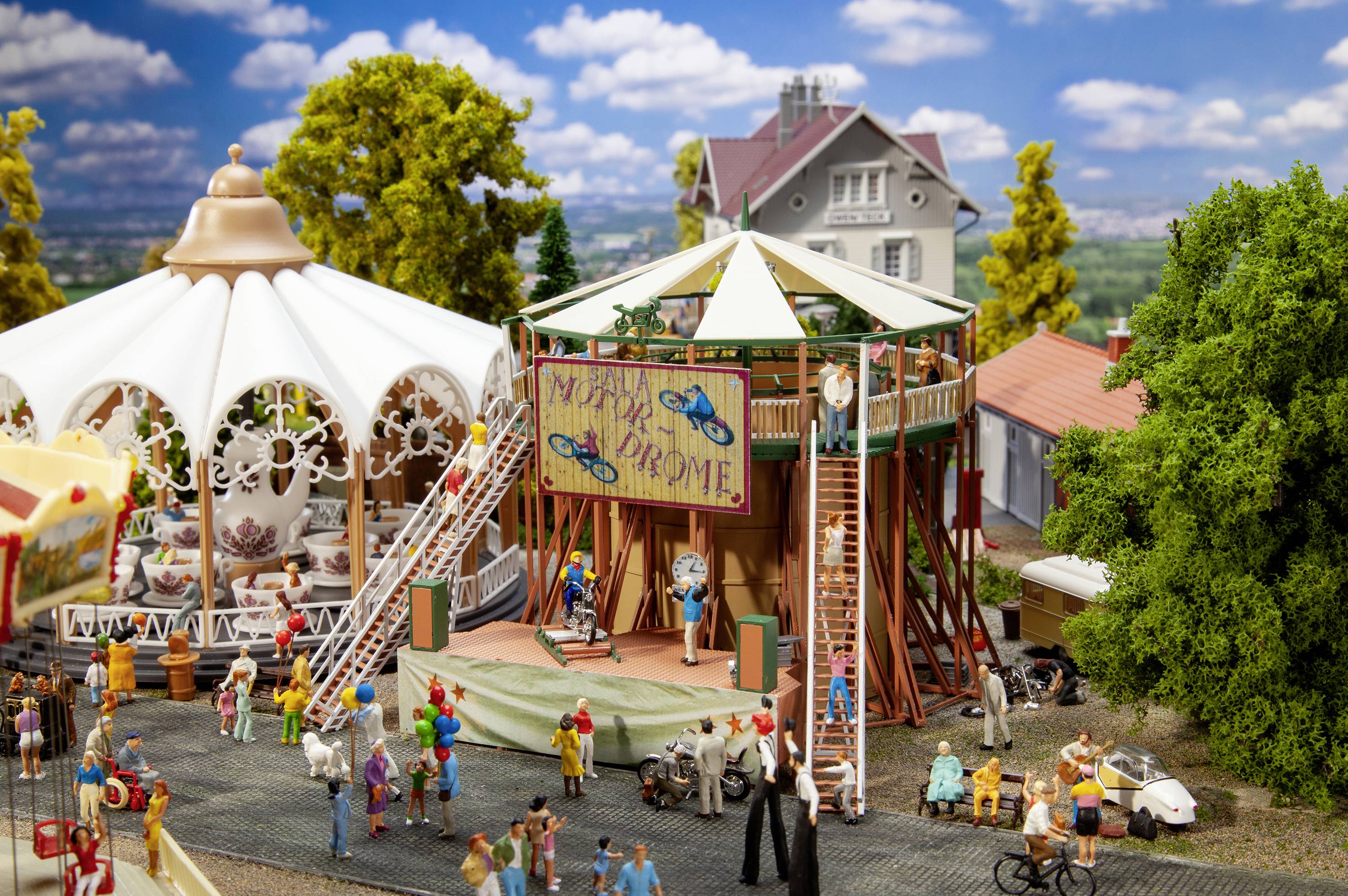 A miniature leisure park with a carousel, stage and people. On the stage is a poster with the inscription 'My Place or My Chrome'.
