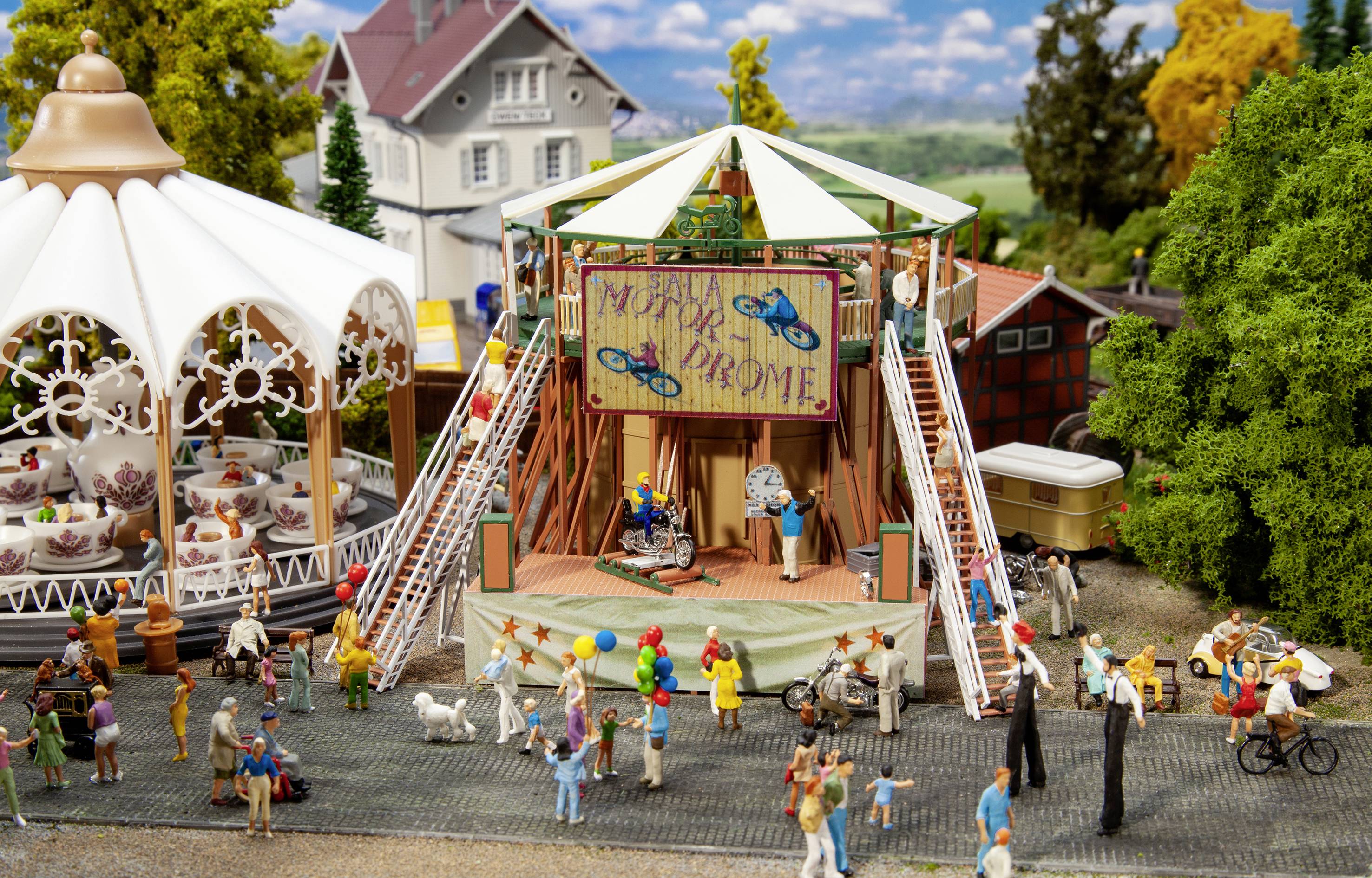 A miniature scene in an amusement park depicts a carousel, a colourful stage with bicycle performers, and many spectators.