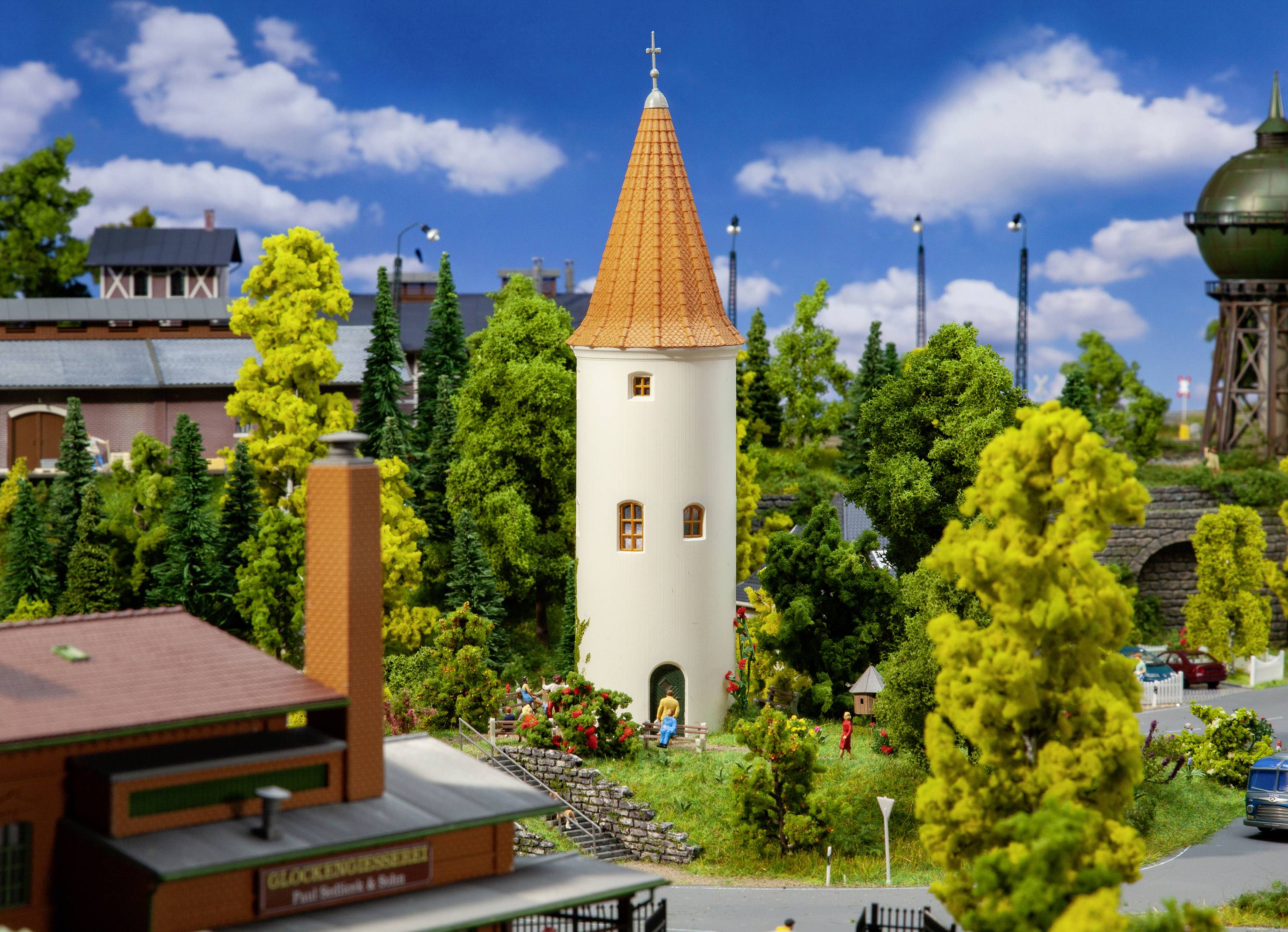 A model village with a white tower in the centre, surrounded by trees and houses, beneath a blue sky with clouds.