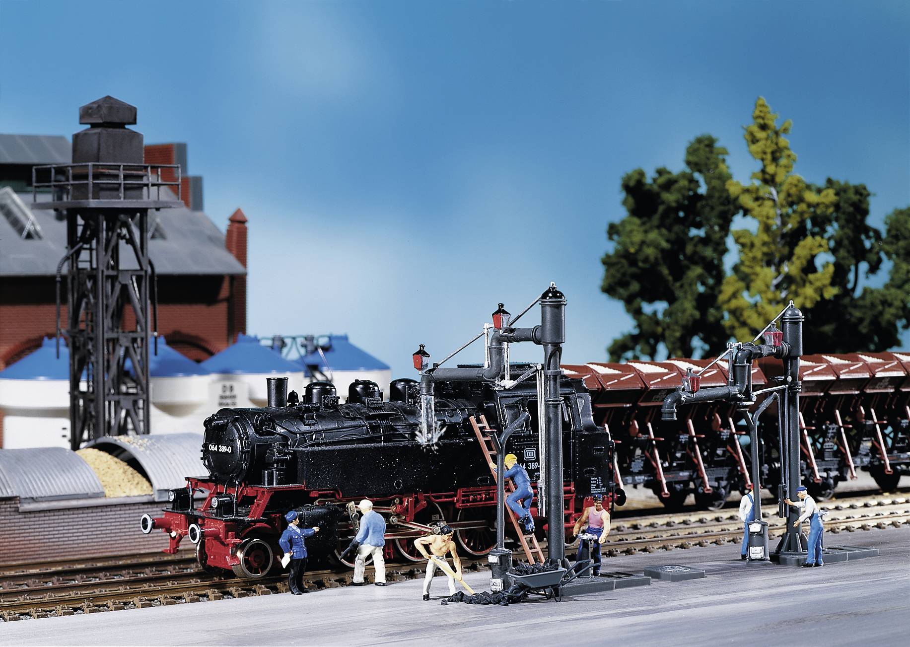 Miniature railway scene with a black steam locomotive, surrounded by small figures working on the tracks. The background shows buildings and trees.